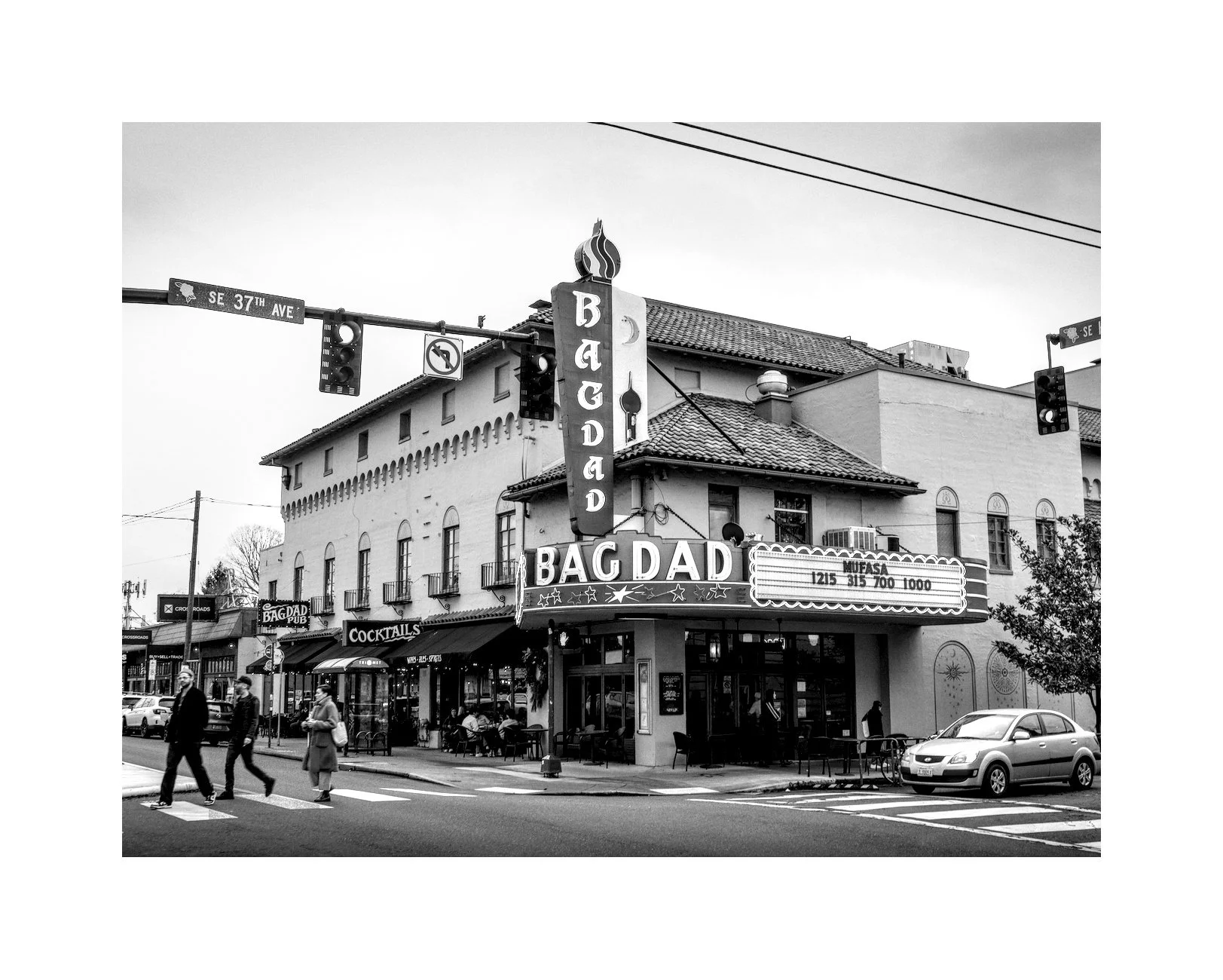 Black and white photo of a street corner with a building that has a marquee sign reading 'BAG DAD'. Pedestrians cross the street, and there are parked cars and storefronts.