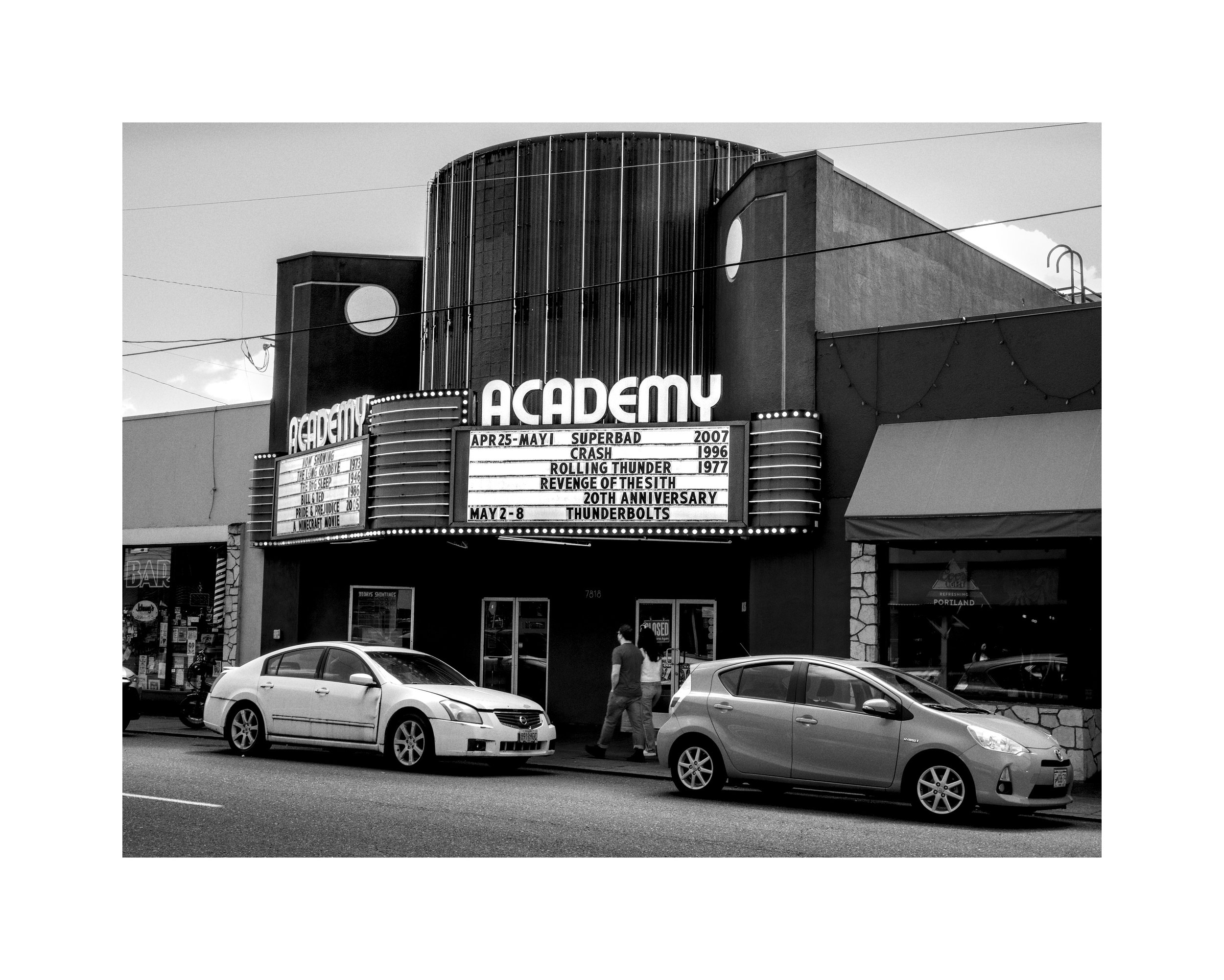 Black and white photo of a theater with the marquee displaying movies and dates, parked cars in front, and a couple walking on the sidewalk.
