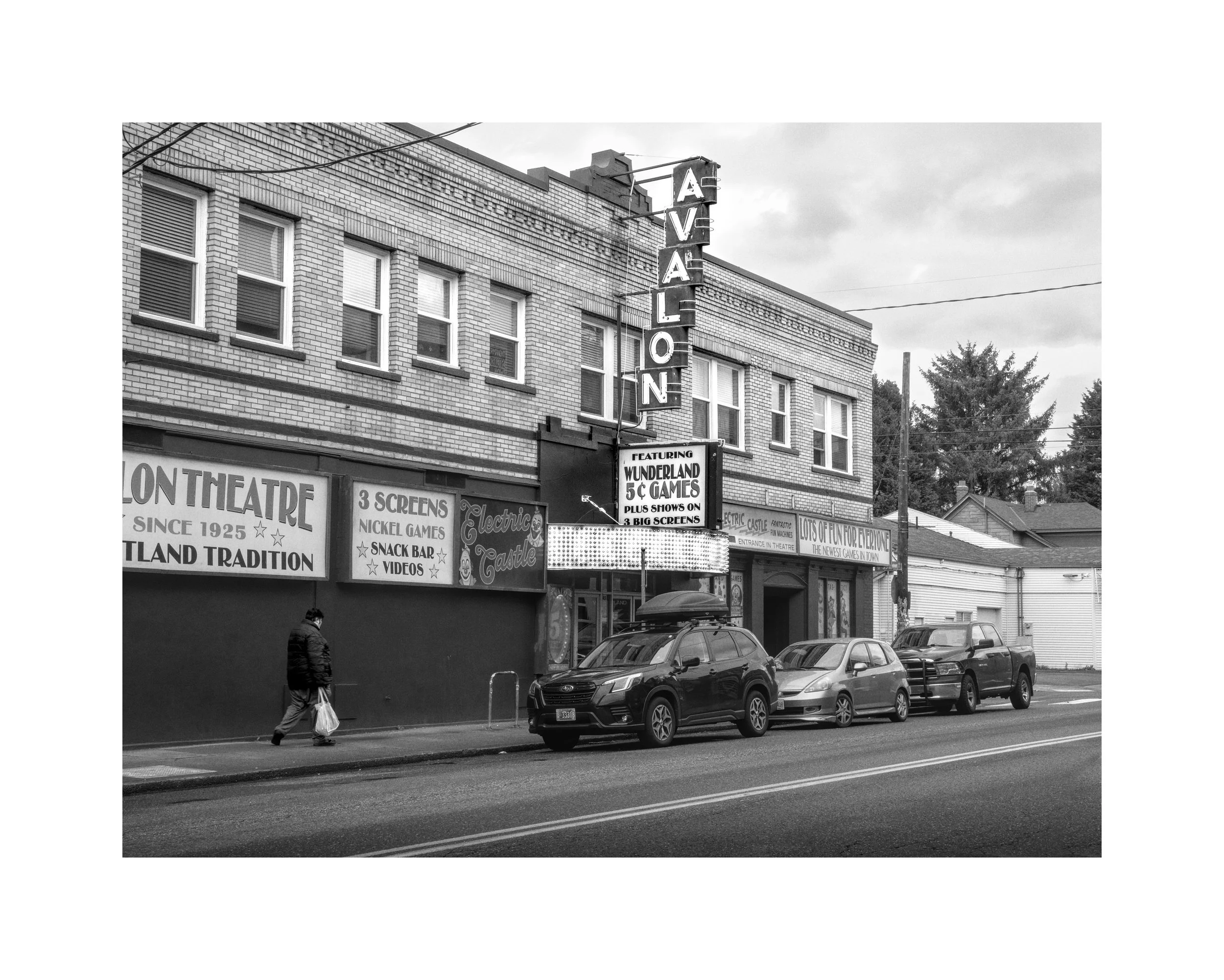 Black and white photo of Avalon Theater with neon sign, parked cars, and a pedestrian walking on the sidewalk.