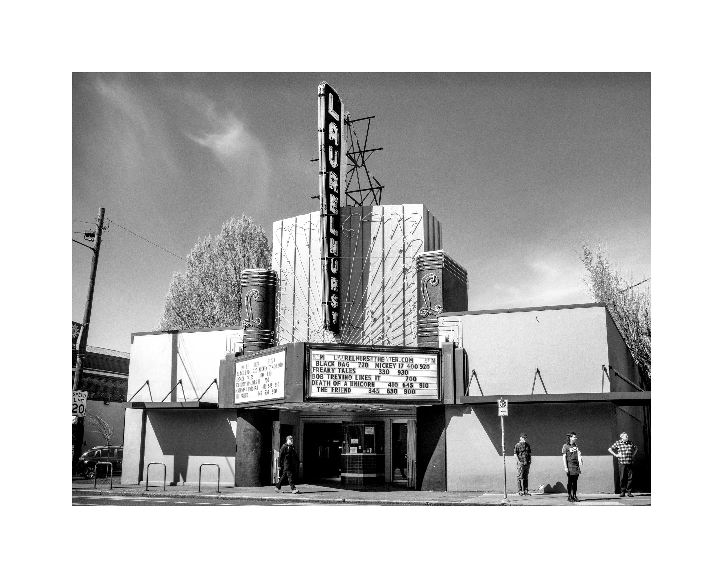 Black and white photo of a vintage movie theater building with a prominent neon sign displaying "FREAKY TALES" and a marquee listing showtimes. There are four people standing in front of the theater, and utility poles and trees are visible in the background.