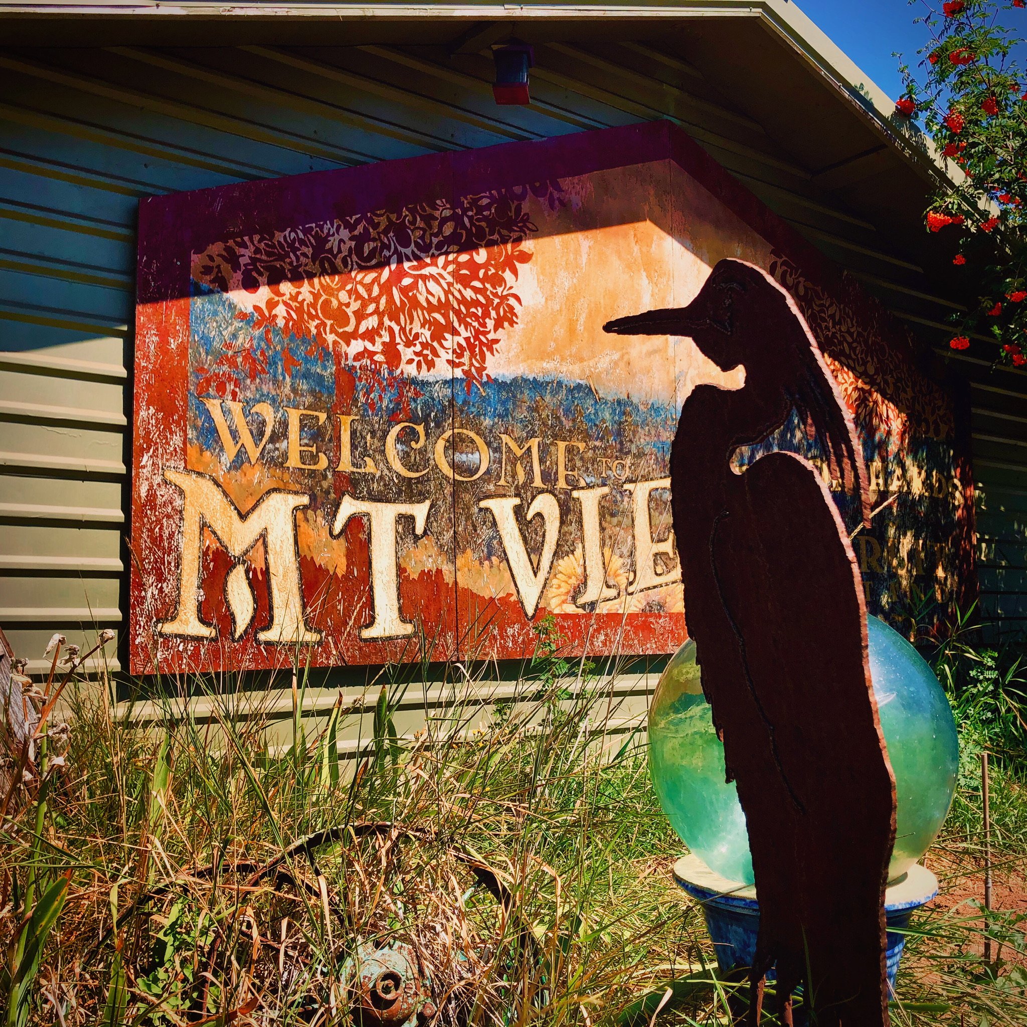 A metal silhouette of a heron standing next to a globe sculpture in front of a wooden sign that says "Welcome to Mt. Vieu" on a house with beige siding and a striped roof. There are plants and a flowering bush around.