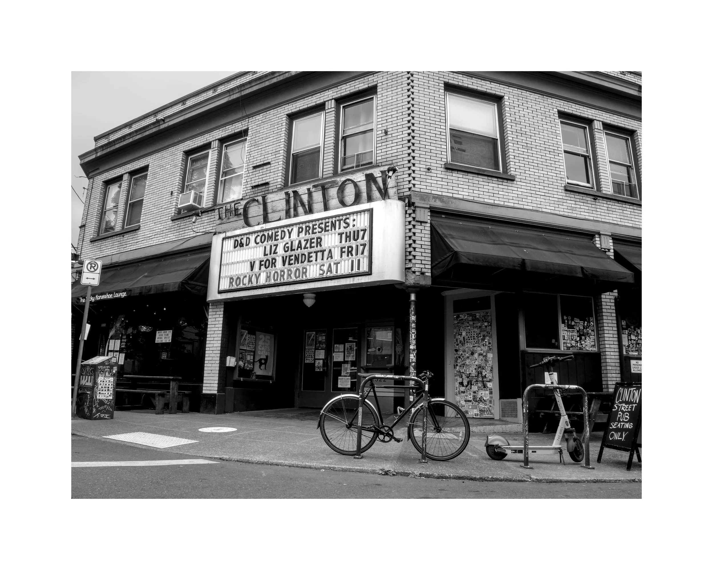 A black and white photograph of a corner building with a theater marquee showing comedy and horror shows. Bicycles and an electric scooter are parked outside, and a sign indicates seating is only for patrons at the Clinton Street Pub.