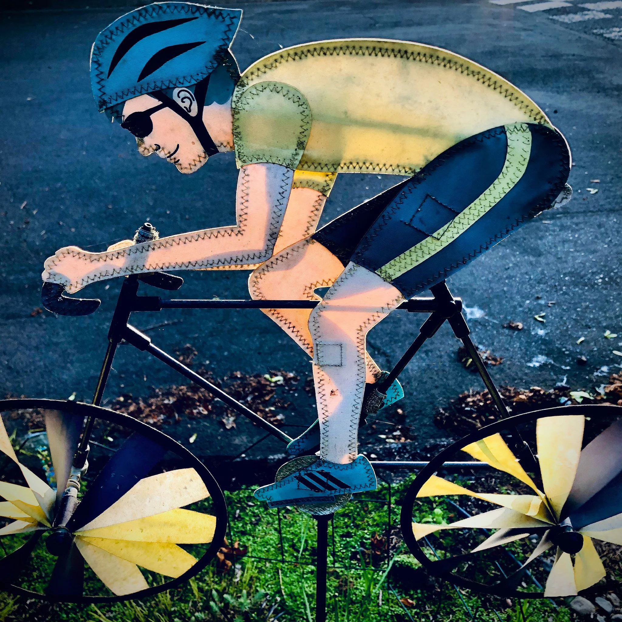Colorful metal sculpture of a cyclist riding a bike, featuring a man wearing sunglasses and a yellow shirt, with a wind-blown hairstyle, set outdoors on a paved surface with grass.