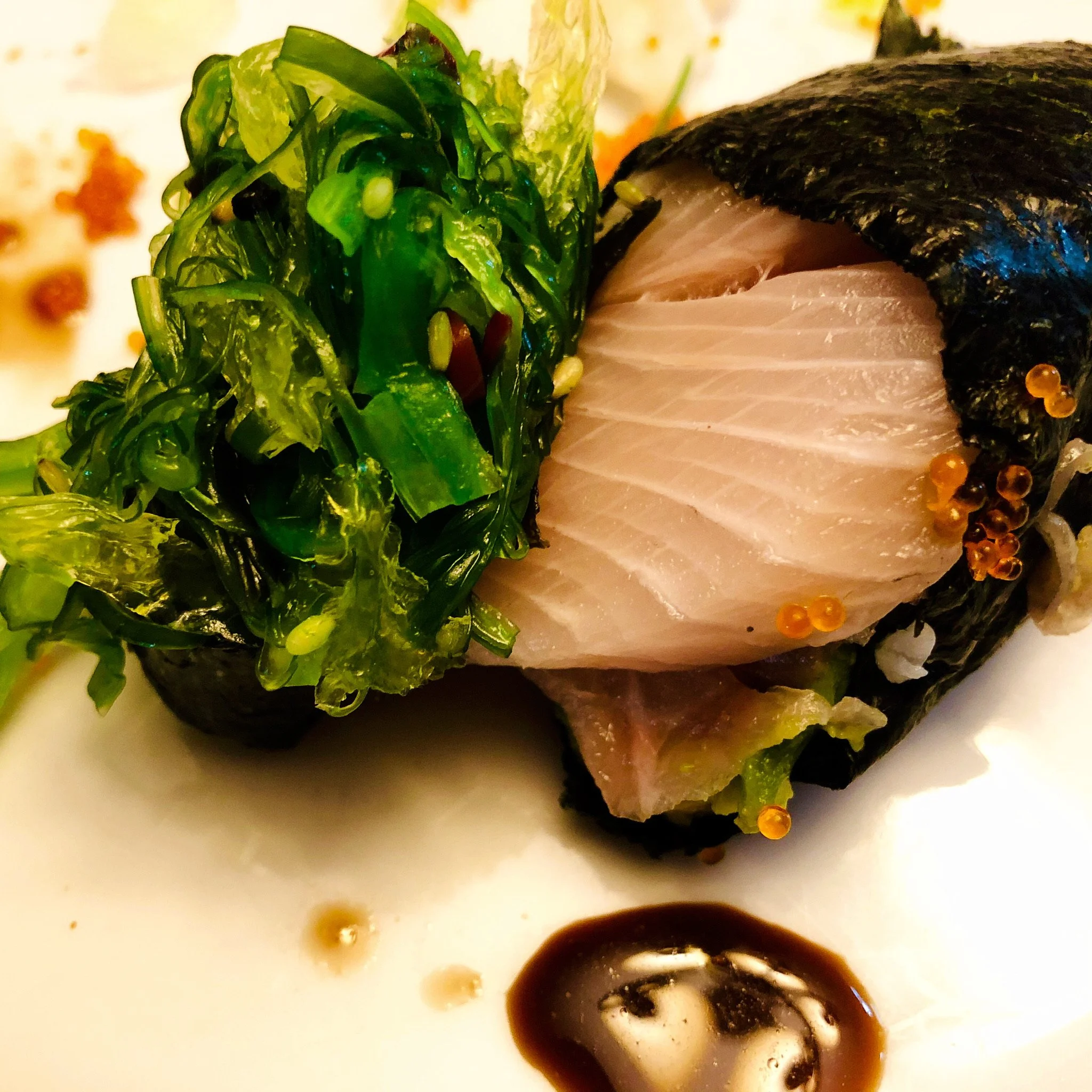 Nigiri sushi with slices of sashimi, seaweed, seaweed salad, and a soy sauce drizzle on a white plate.