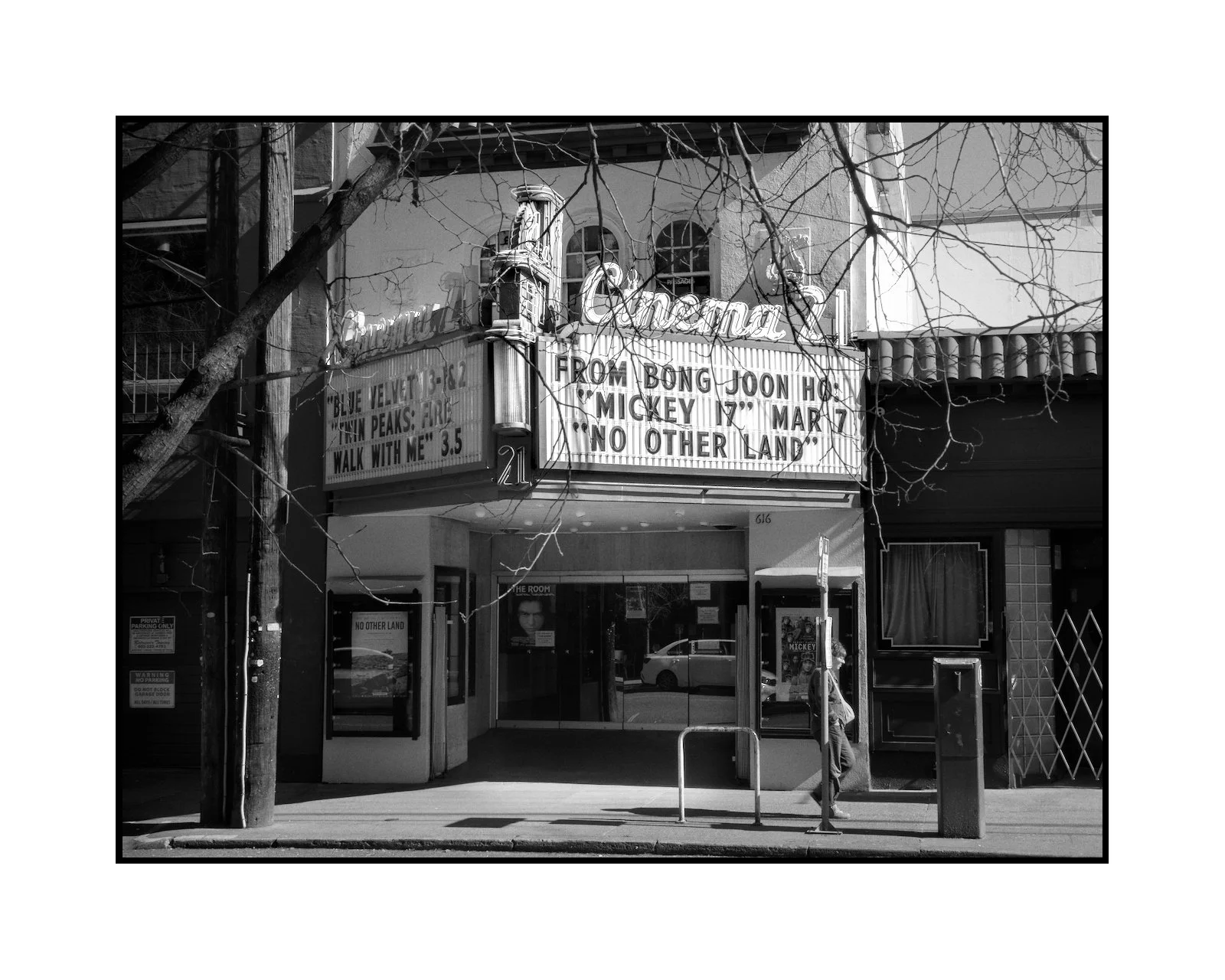 The exterior of a theater with a marquee advertising a performance about Mickey Mouse on March 7, with a quote, and a person walking past on the sidewalk.