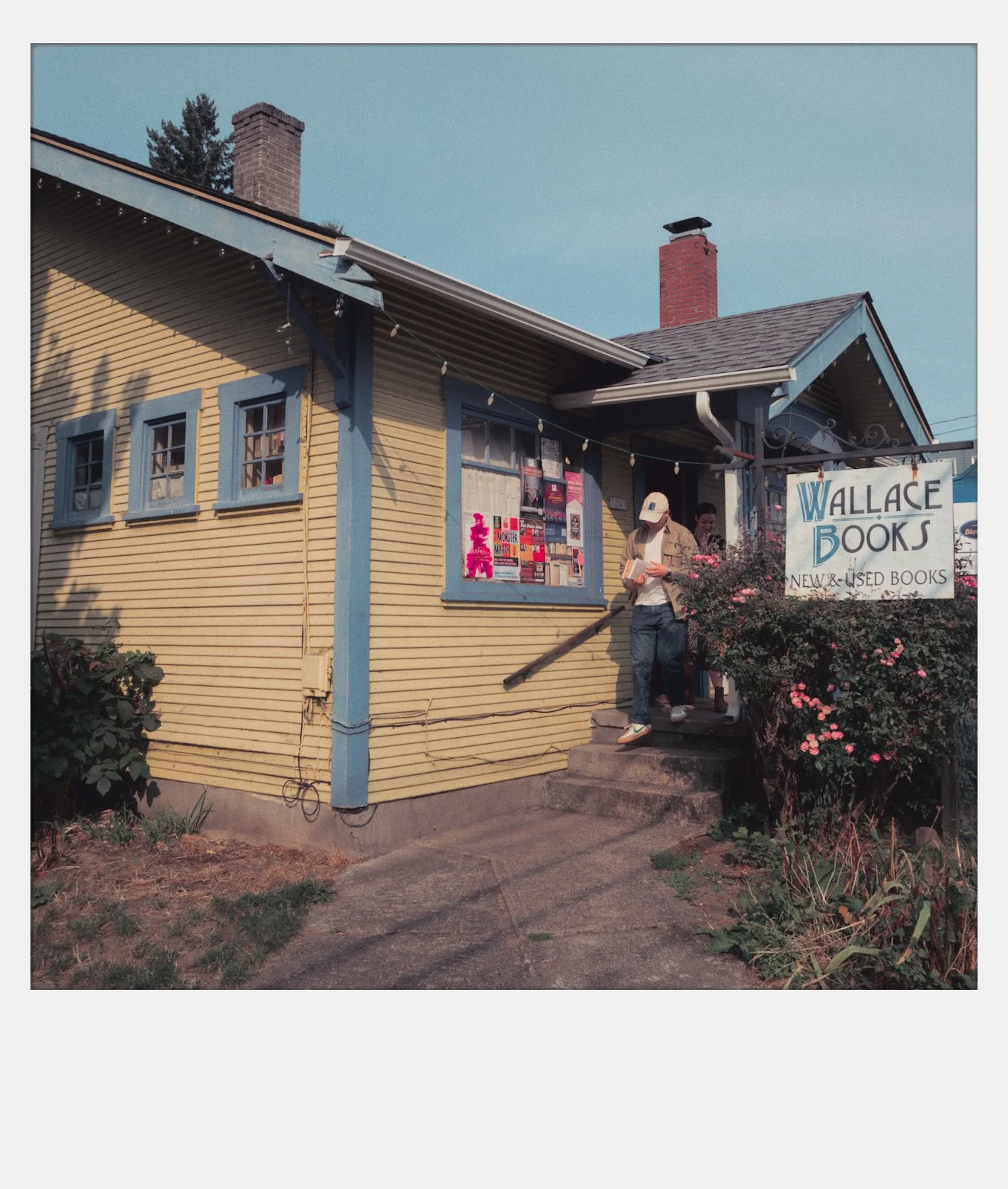 A yellow house with blue window frames and a sign that reads 'Wallace Books, New & Used Books.' Two people are walking out of the house, holding books, with a bush of pink flowers nearby.