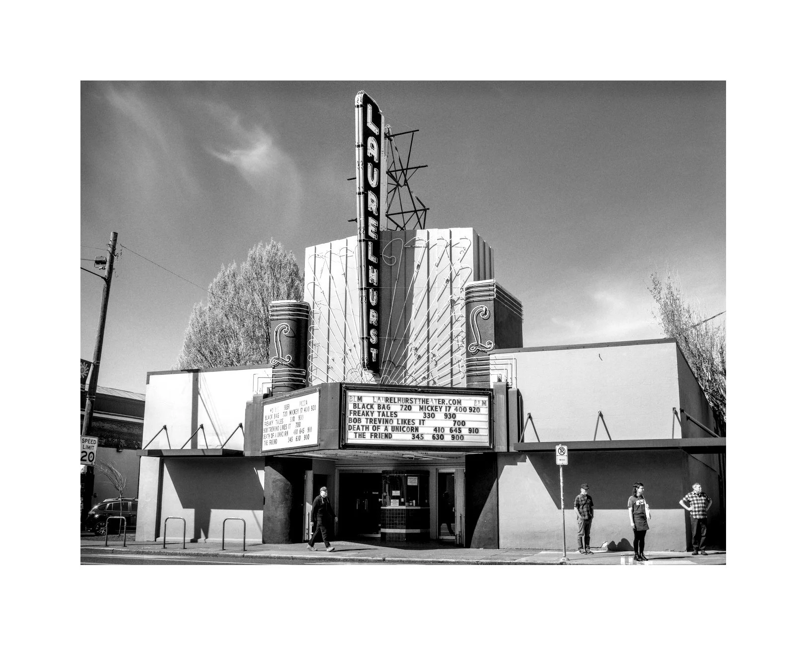 Black and white photo of a vintage movie theater with marquee sign, four people standing outside, and tall trees in the background.