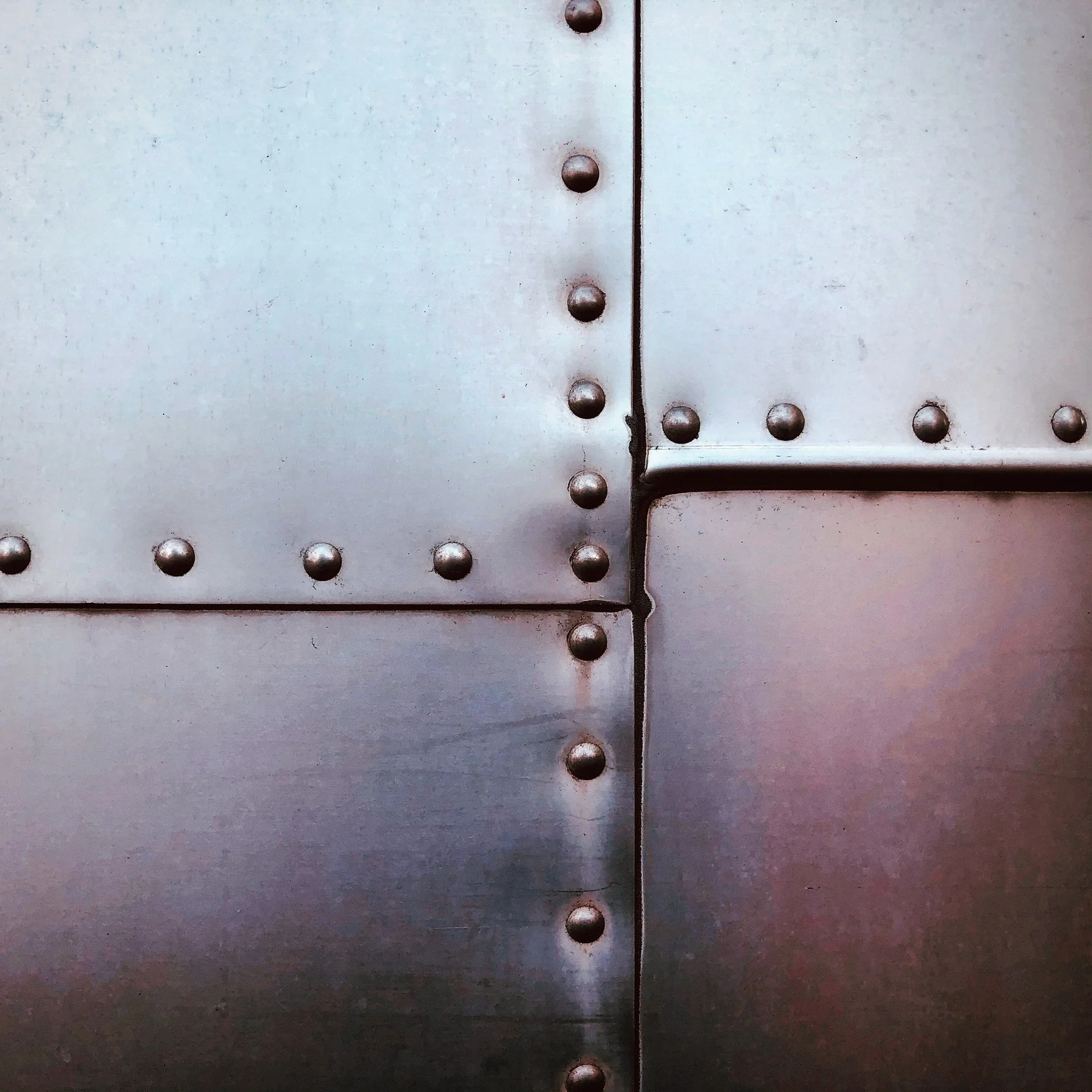 Close-up of a metal surface with visible rivets and a seam, featuring a vertical and a horizontal panel.