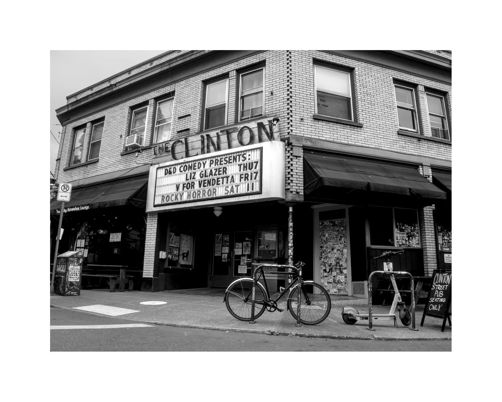 Black and white photo of a corner building with a theater marquee, bicycles, and a scooter on the sidewalk, and a sign indicating only street pub seating.