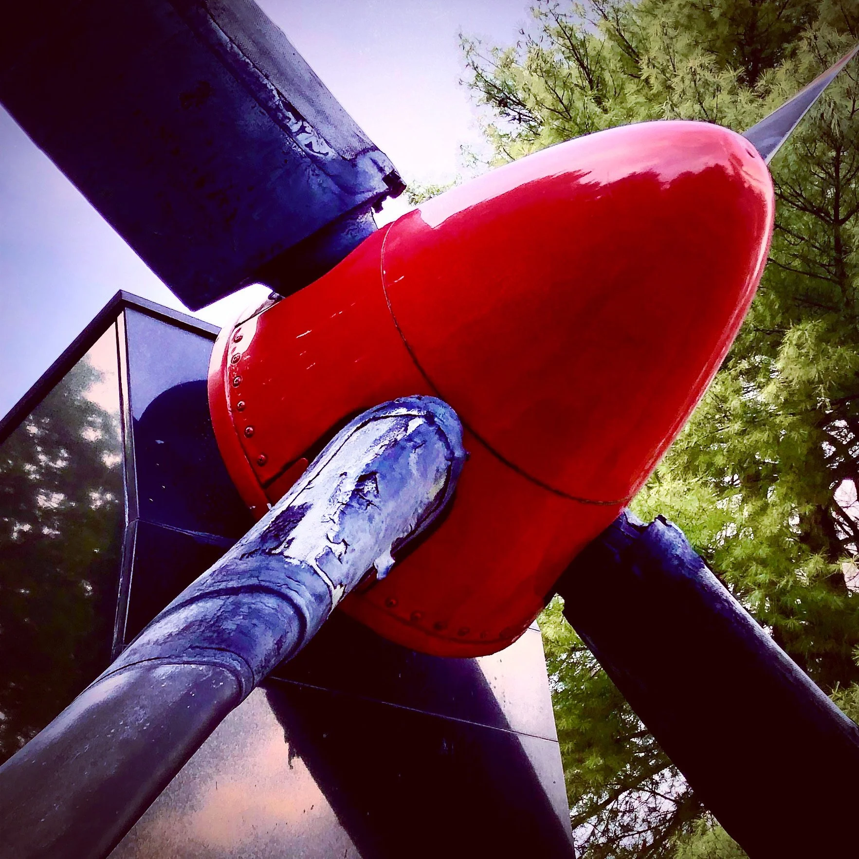 Close-up of the front section of a red rocket sculpture mounted on black supports with trees in the background.