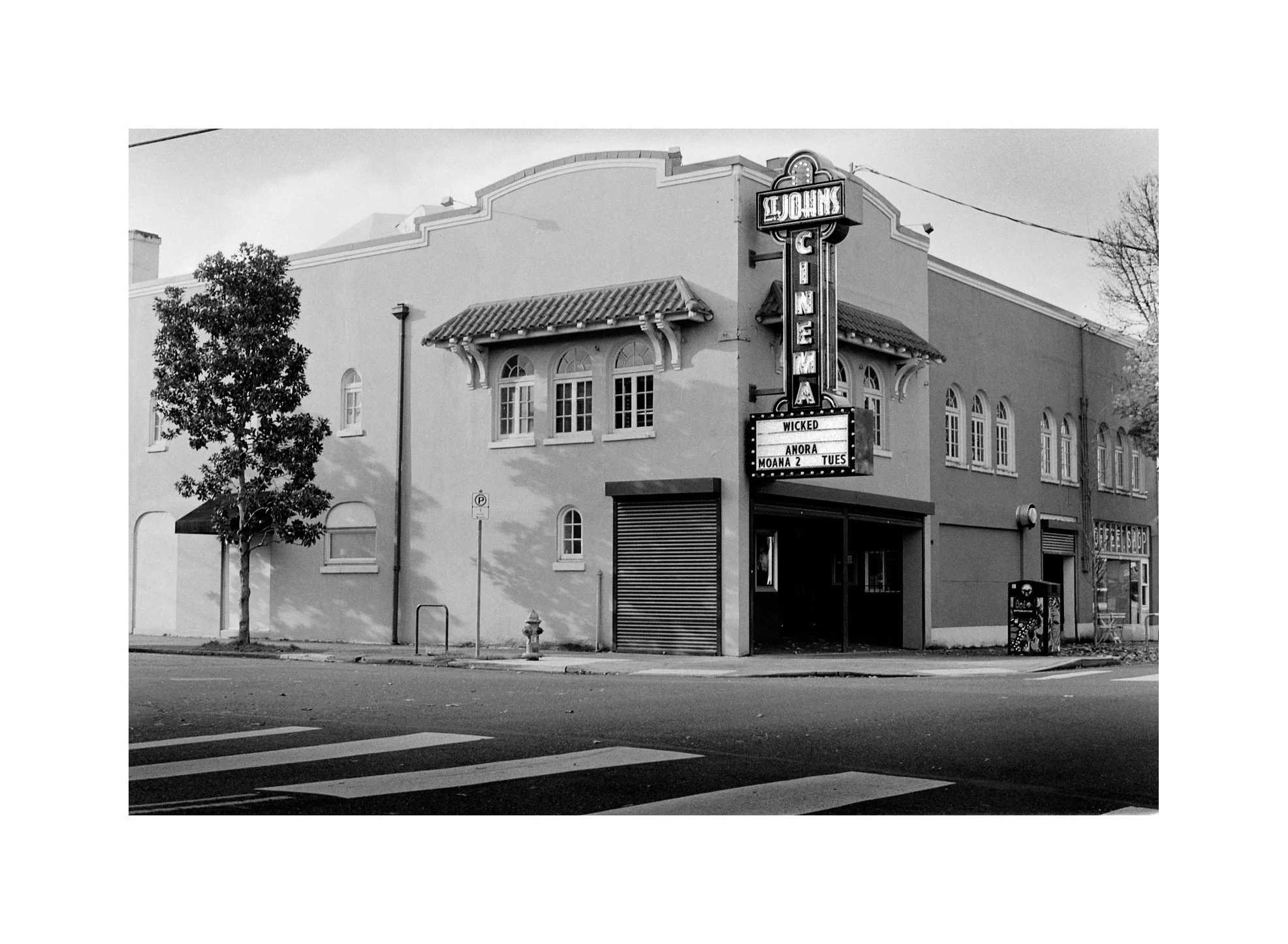 Black and white photo of a vintage theater with a lit vertical sign reading 'S. John Cinema' and a marquee sign displaying movie titles.
