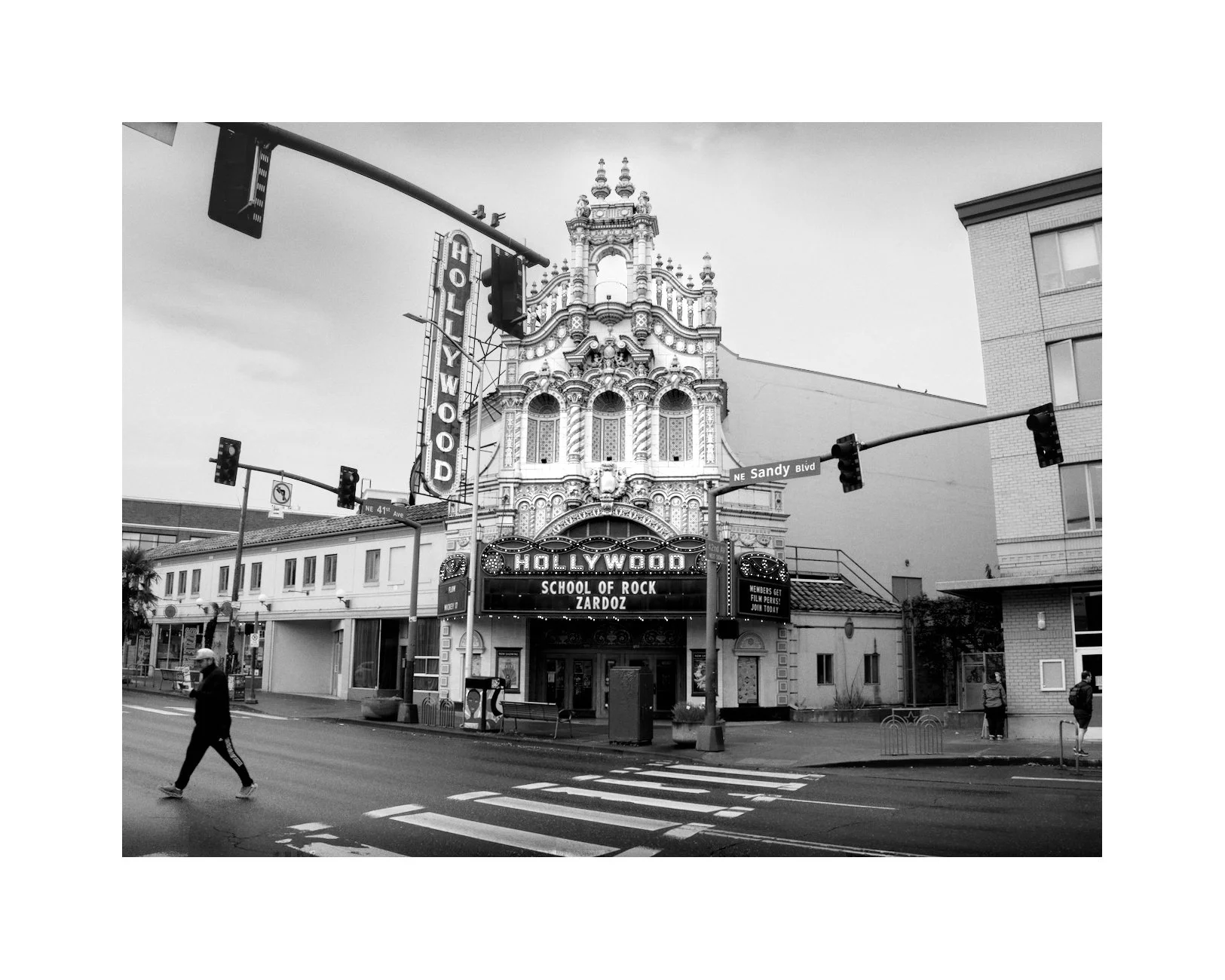A black and white photo of the Hollywood Theatre in Portland, Oregon, with an ornate facade and marquee reading "Hollywood School of Rock Zardoz". Pedestrians cross the street, and traffic lights and street signs are visible.
