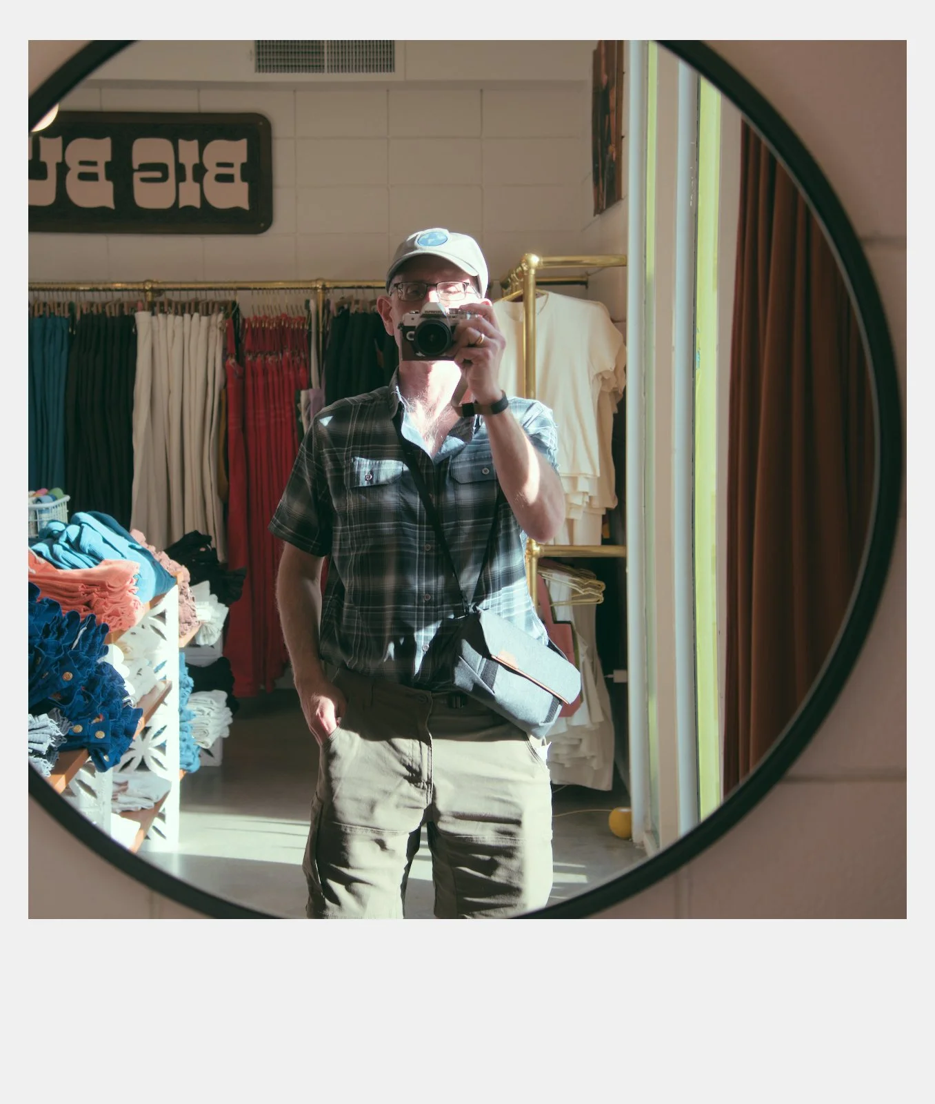 A man taking a selfie in a fitting room mirror, wearing a plaid shirt, beige pants, a white baseball cap, and glasses, with a camera around his neck and a gray bag on his chest, with colorful clothing and curtains in the background.