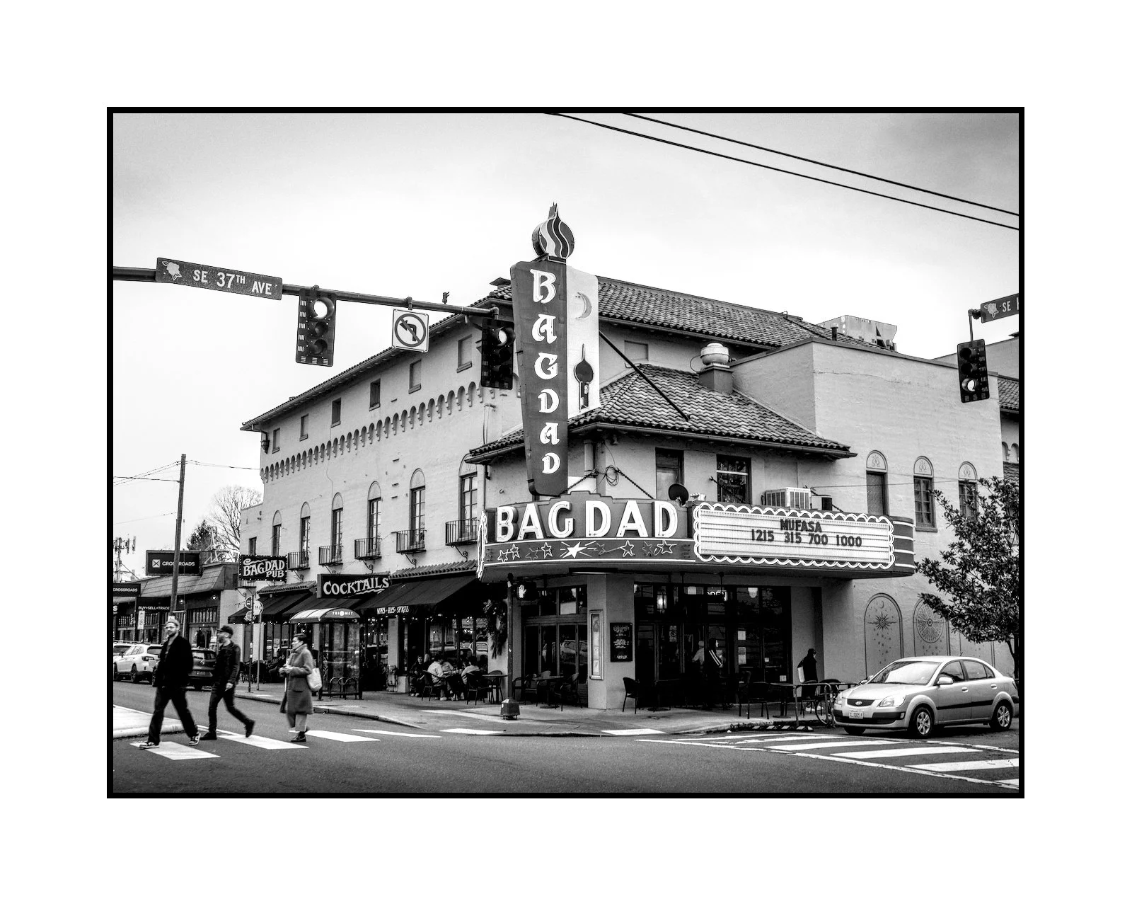 A black and white photo of a corner building with a sign for a tavern called Bagdad Pub, located on a street corner with pedestrians crossing the street and a parked car in front.