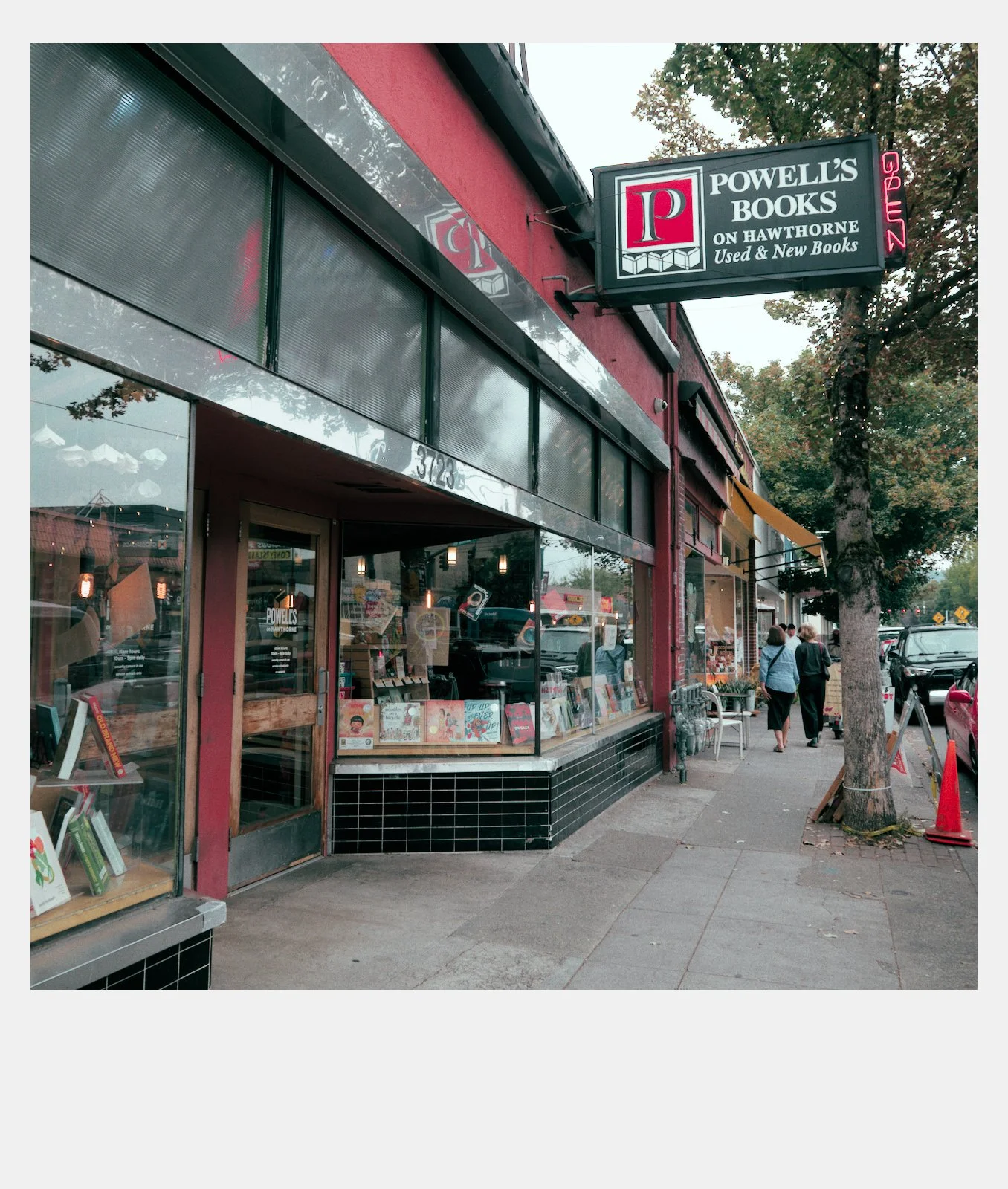 Exterior view of Powell's Books store on Hawthorne, with a sign indicating it sells used and new books, and a neon 'open' sign. People walking along the sidewalk.
