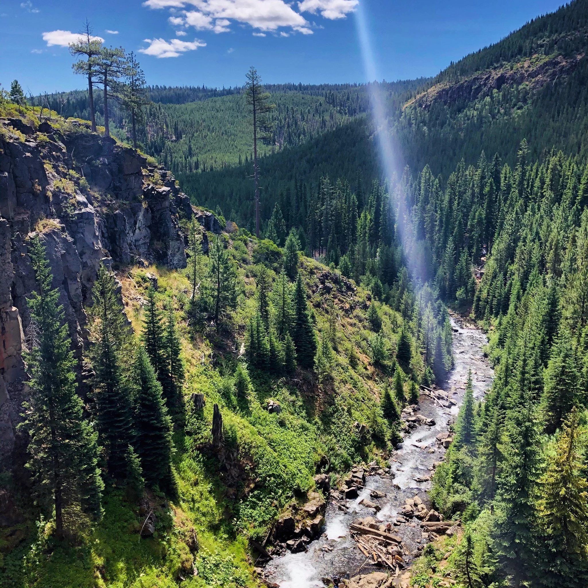 A lush green mountainous landscape with a rocky cliff on the left, dense evergreen trees, a river flowing through the valley, and a sunbeam streaking through a partly cloudy sky.