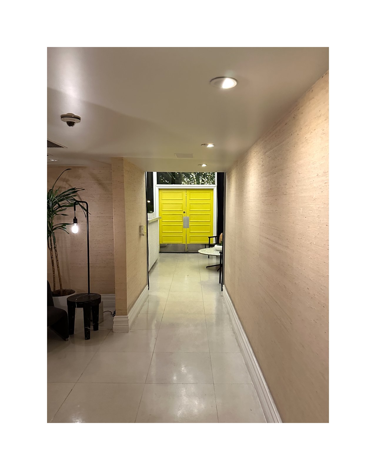 Interior hallway with cream-colored walls, tiled floor, ceiling lights, and a bright yellow door at the end, with some furniture and a potted plant on the left side.