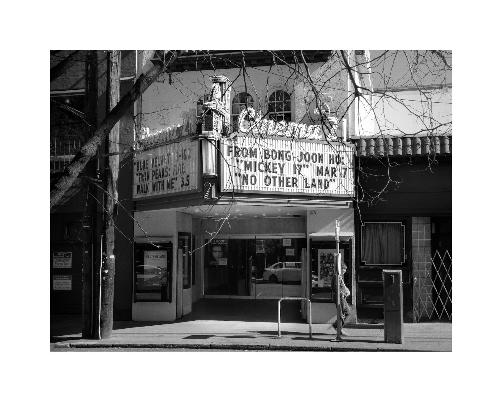Black and white photo of a movie theater with a marquee announcing showings, including 'Blue Velvet,' 'Twin Peaks: Fire Walk with Me,' and a special event on March 7th. A person walks past the theater entrance on the sidewalk, with trees and building