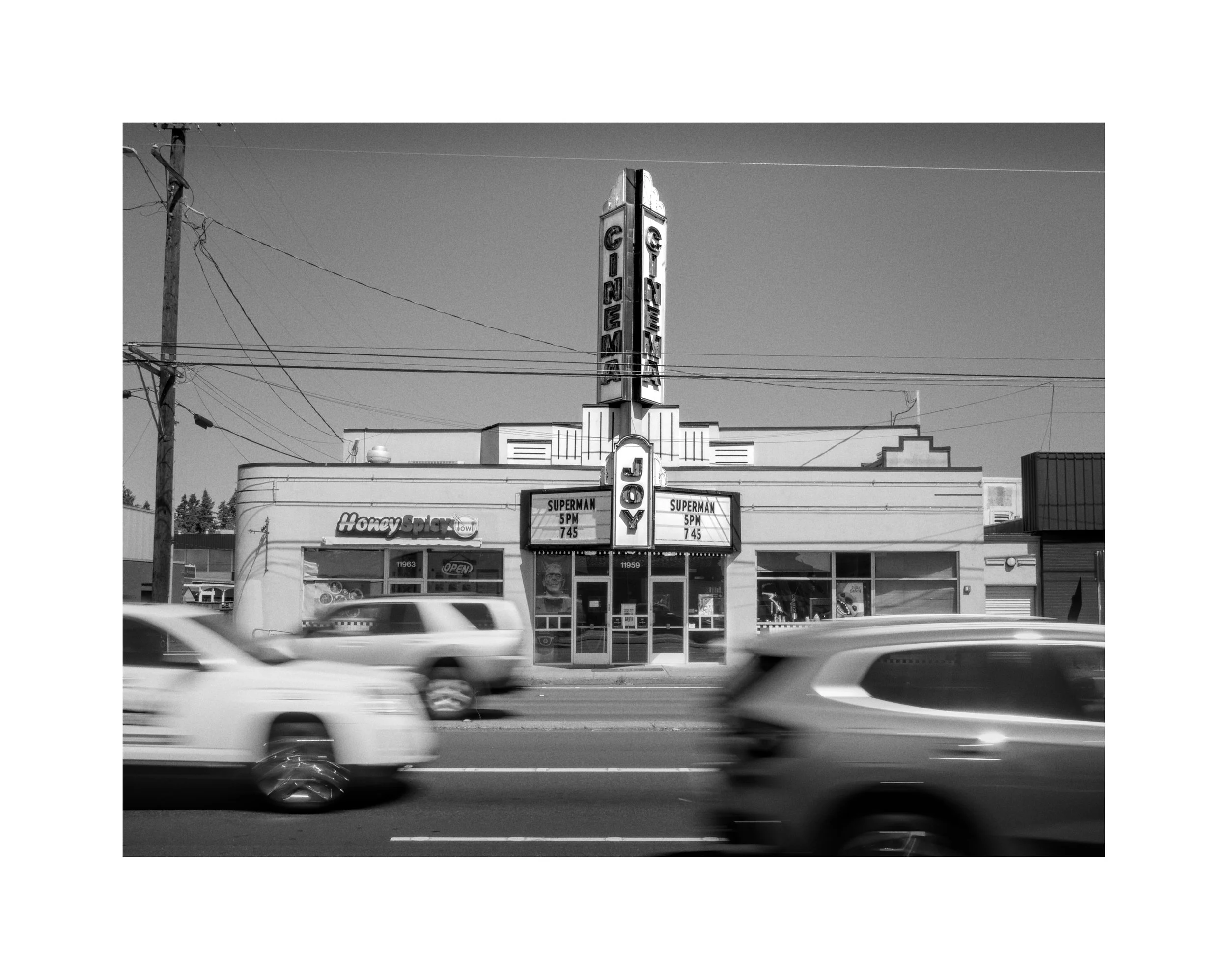 Black and white photo of a vintage movie theater with vertical sign reading 'CINEMA' and marquee advertising a Superman showing at 5 pm, with blurred cars passing in front.