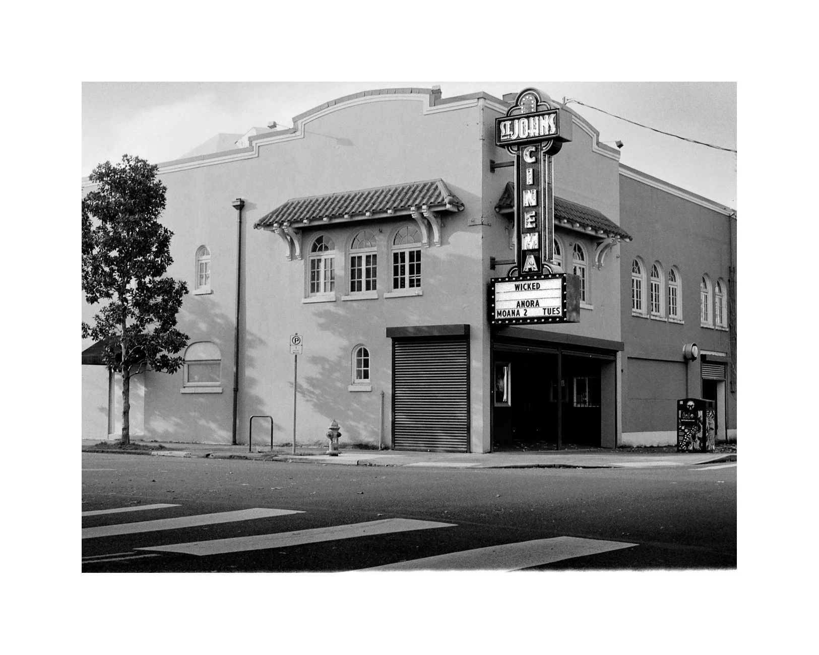 Black and white photo of an old theater building with a vertical marquee sign displaying the word 'Screen' and a smaller sign with movie titles, including 'Wicked,' 'Anora,' and 'Moana 2,' on a city street.