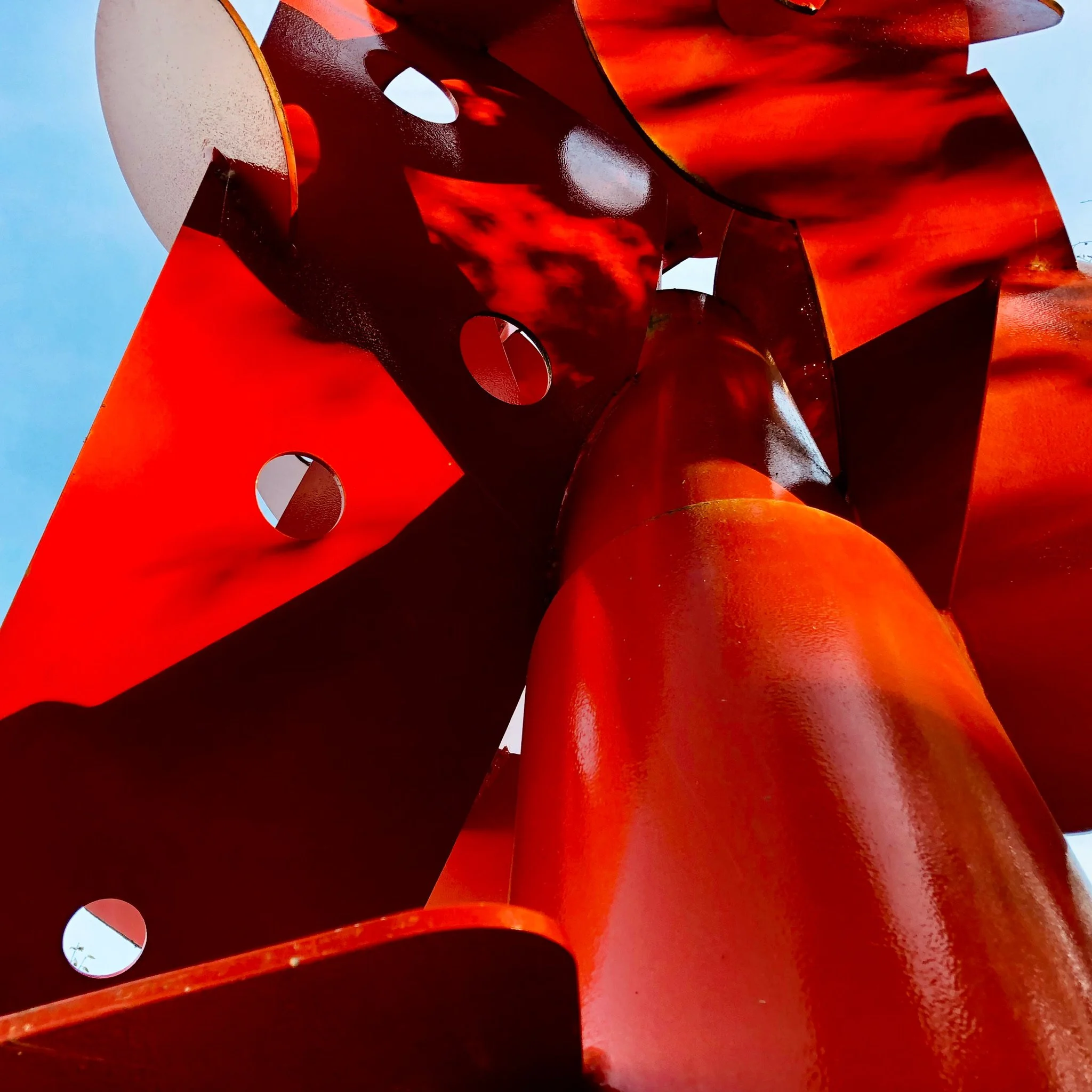 Close-up of a large abstract red metal sculpture with circular cutouts, viewed from below against a blue sky.