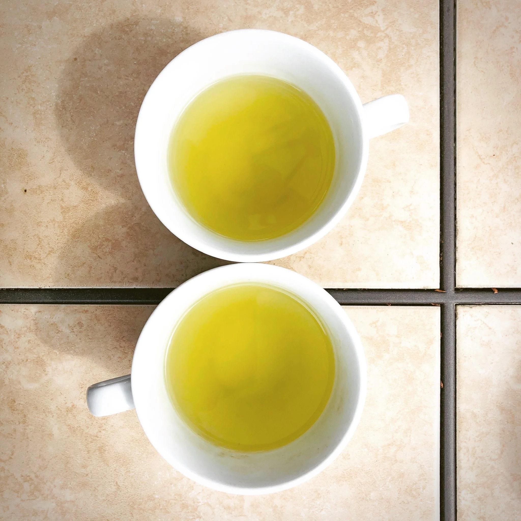 Two white mugs filled with green tea placed on a tiled surface.