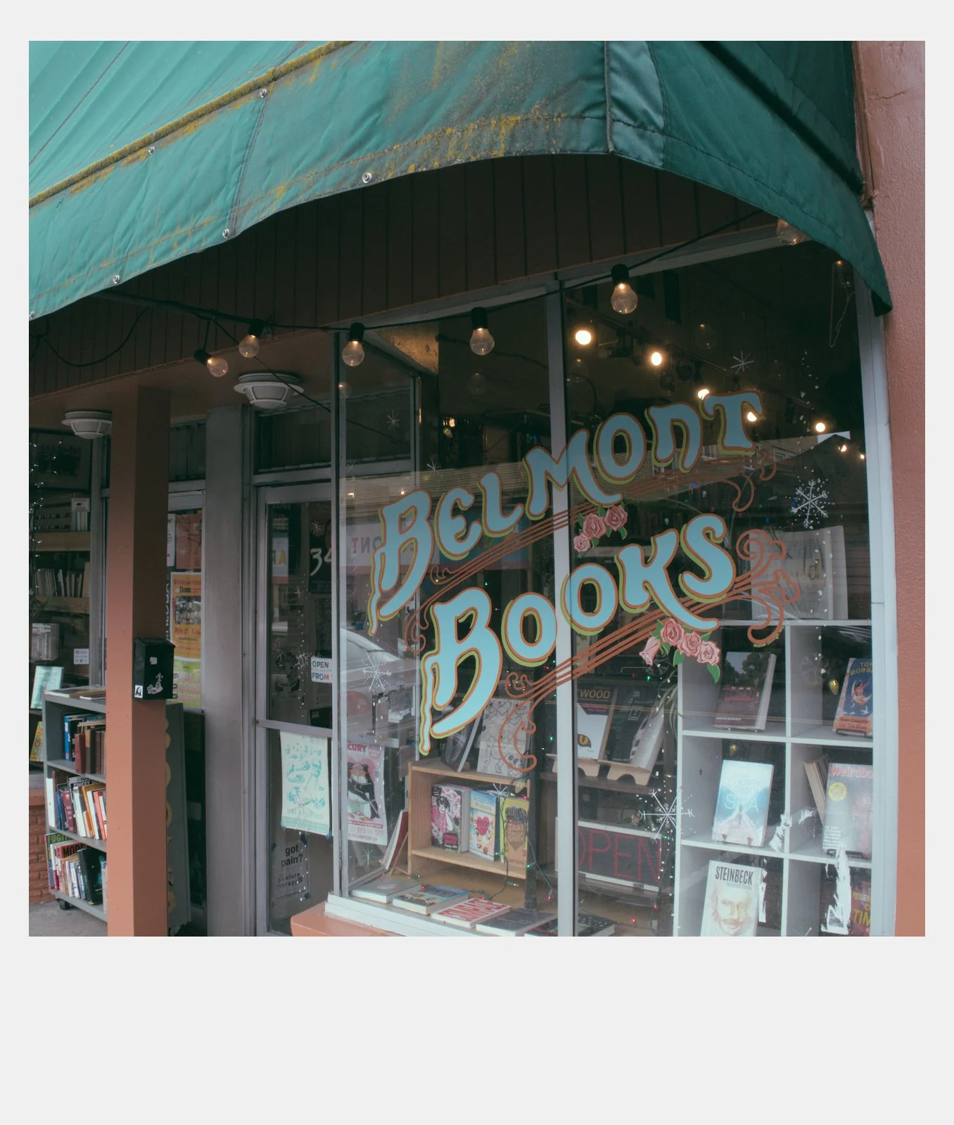 Bookstore storefront with large window display reading 'Belmont Books' decorated with pink roses and snowflakes, inside are books, a small table, and an open sign.