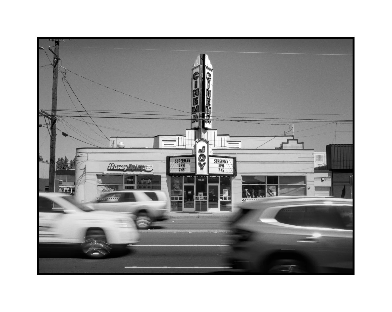 Black and white photo of a vintage cinema building with a vertical sign reading 'CINEMAS' and 'MOVIE,' cars passing by in the foreground, and a small shop named 'Honey Sply' on the left.