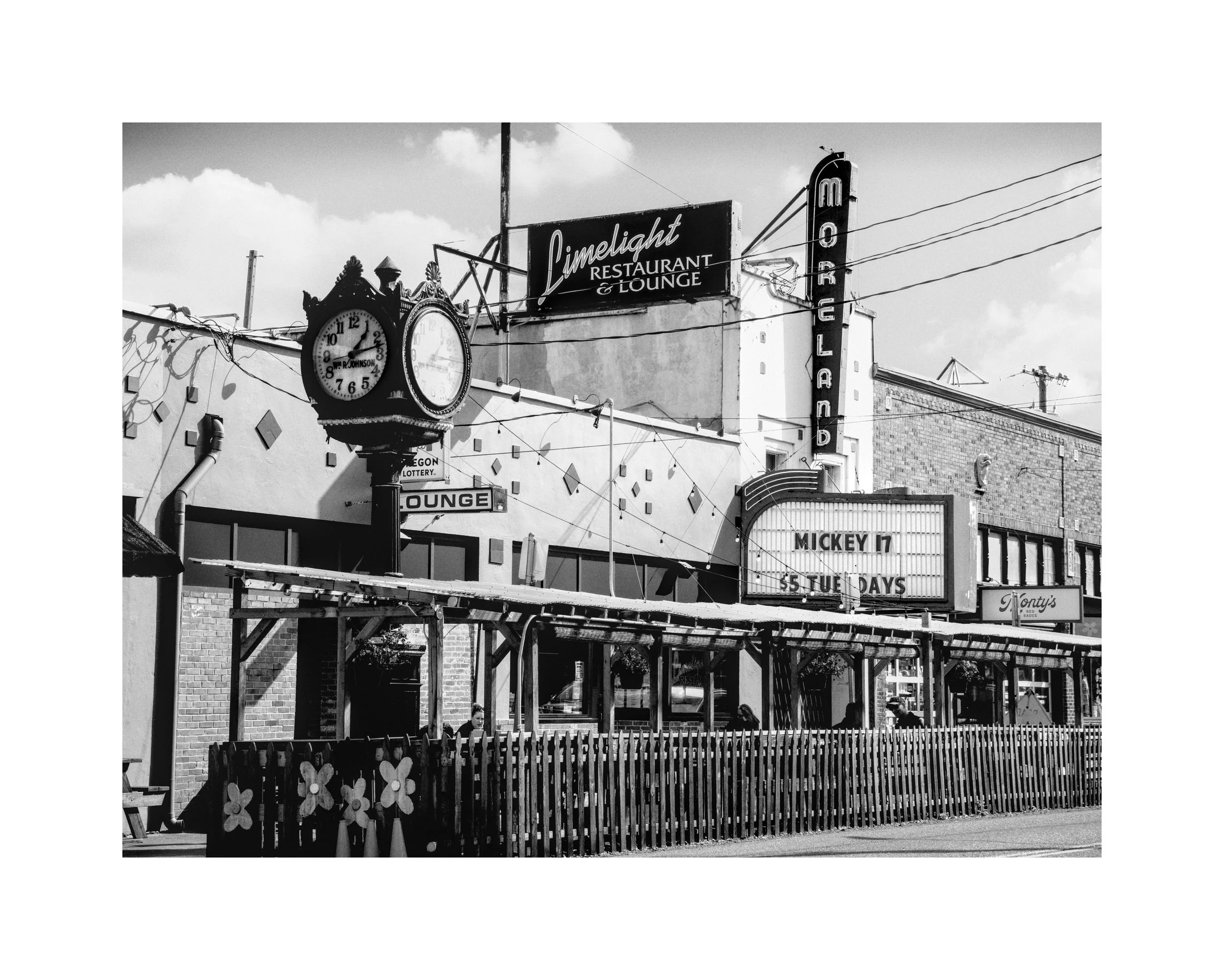 Black and white photo of a street scene with vintage signs, a large clock, and a storefront for a restaurant and lounge, with pedestrians walking by.