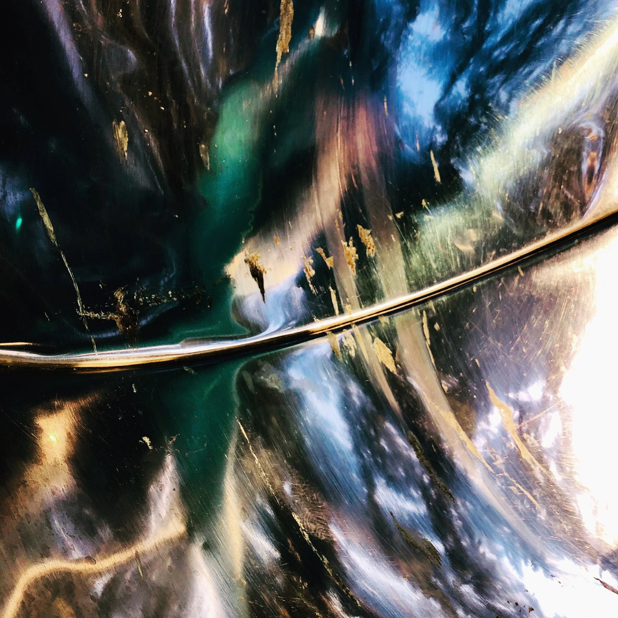Close-up of a shiny, reflective, metallic surface with streaks of black, green, blue, and gold colors, showing a curved line or seam across the surface.