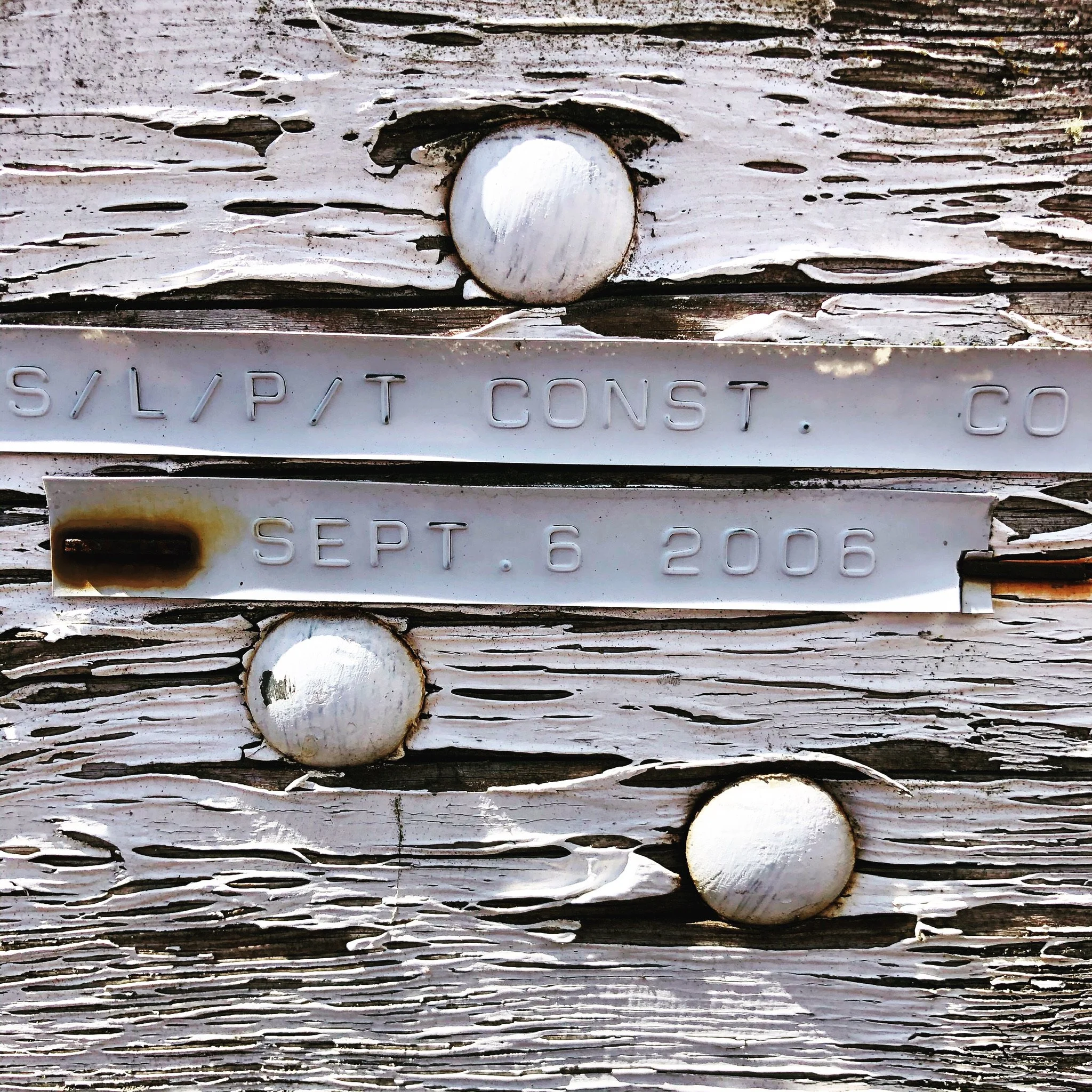 Close-up of a weathered wooden surface with three round white nails and two metal plates, one with faint inscriptions and the other with the date 'Sept. 6, 2006' inscribed.