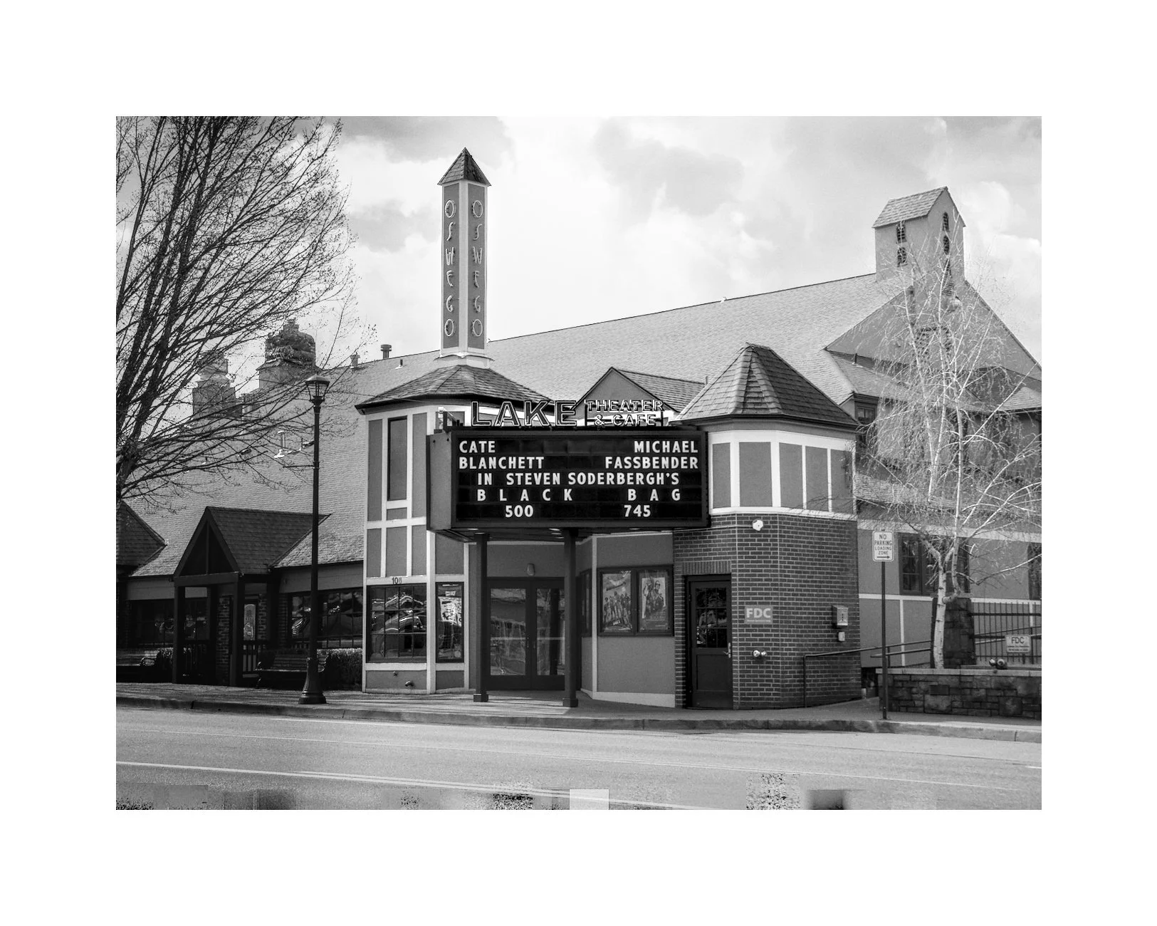 Black and white photo of a theater building with a marquee and vertical sign that reads "Oaks Oscar." The marquee displays show titles and times, with the words "Lake Theater" on the top of the building.