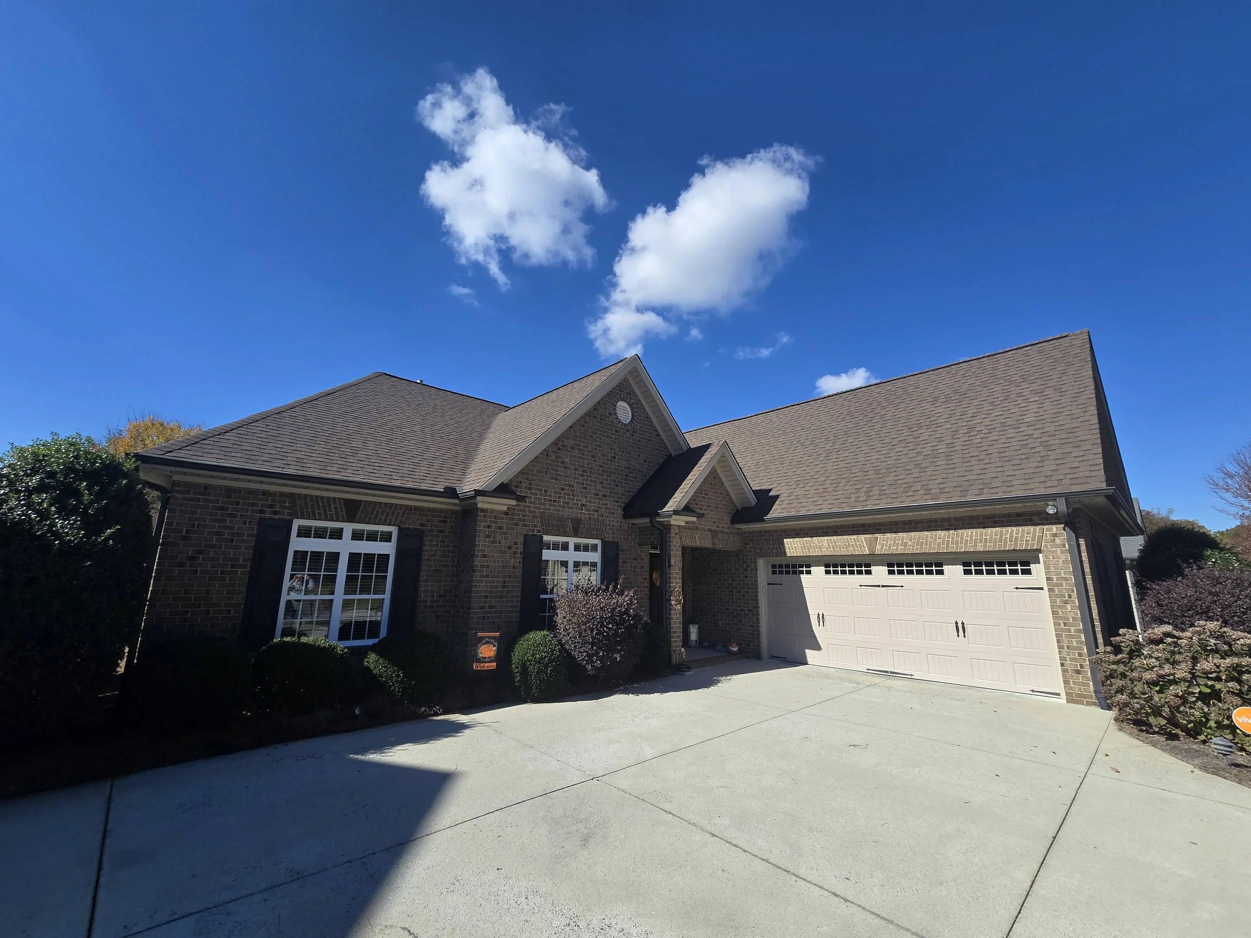 A residential house with a brick exterior, two-car garage, and front yard. The house has dark shutters on the windows, and the roof is sloped with brown shingles. There are trees and bushes around the house. The sky is clear with a few white clouds.