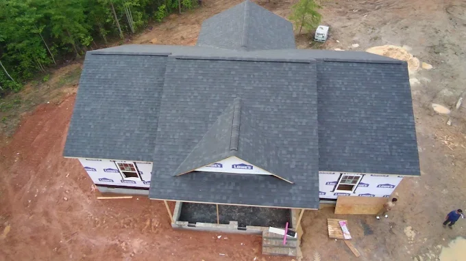 An aerial view of a house under construction with a gray shingle roof, surrounded by construction site dirt and some trees in the background.