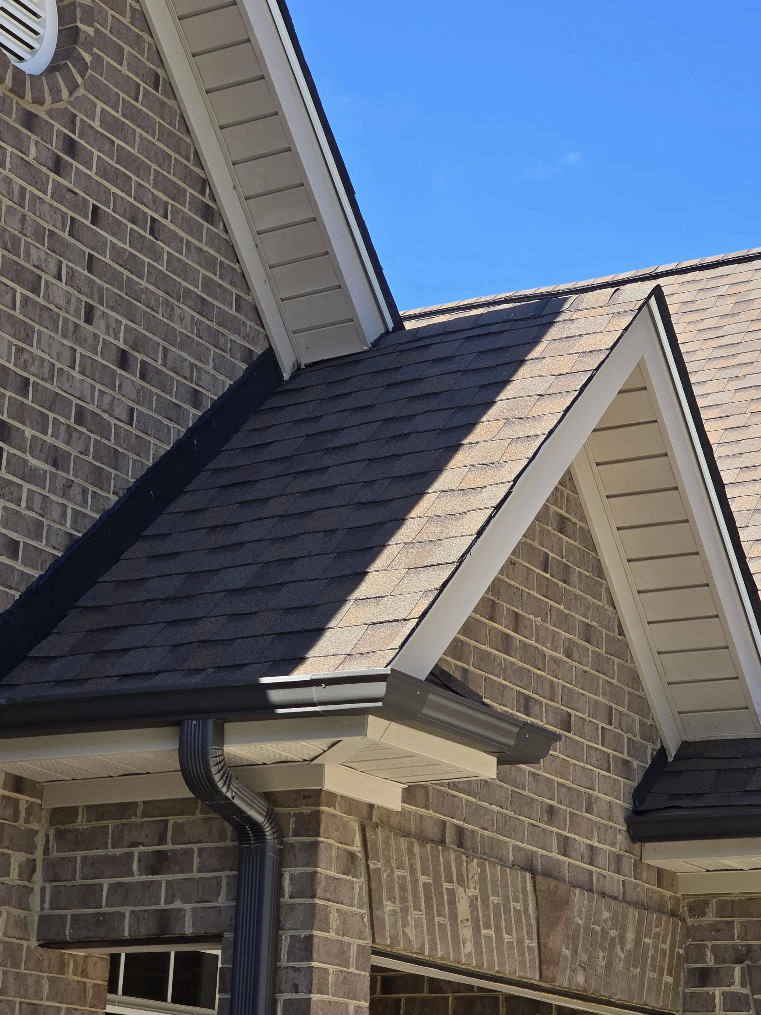Close-up view of the corner of a house showing the brick wall, roof, and gutter system with black downspouts, under a clear blue sky.