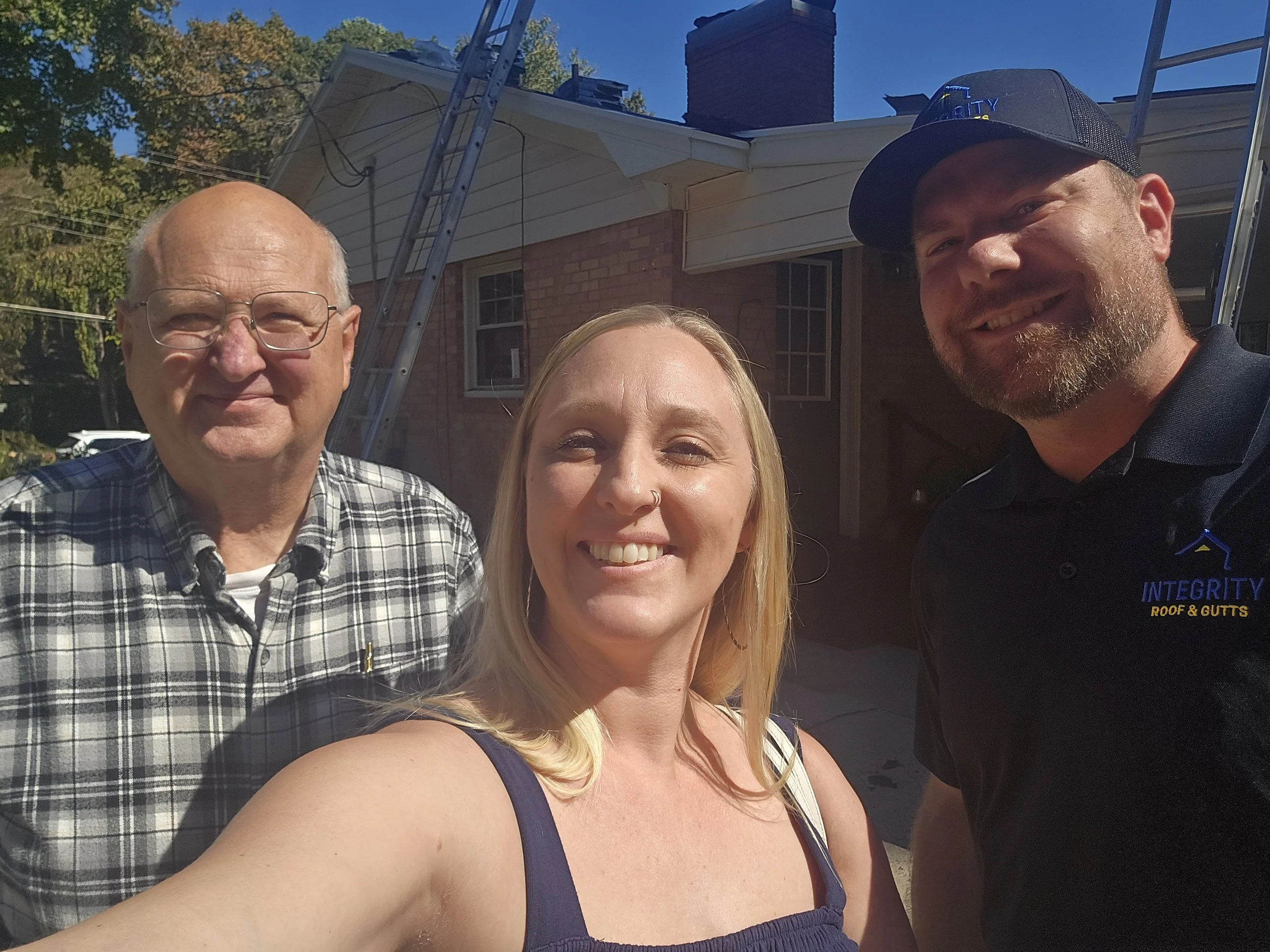 Three smiling people taking a selfie outdoors in front of a house with a ladder and chimney, one woman and two men, one wearing a black cap with the words 'Integrity Roof & Gutters'.
