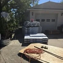 Pallets of stacked bricks on a driveway near a garage, with a coil of orange extension cord and a hammer in the foreground.