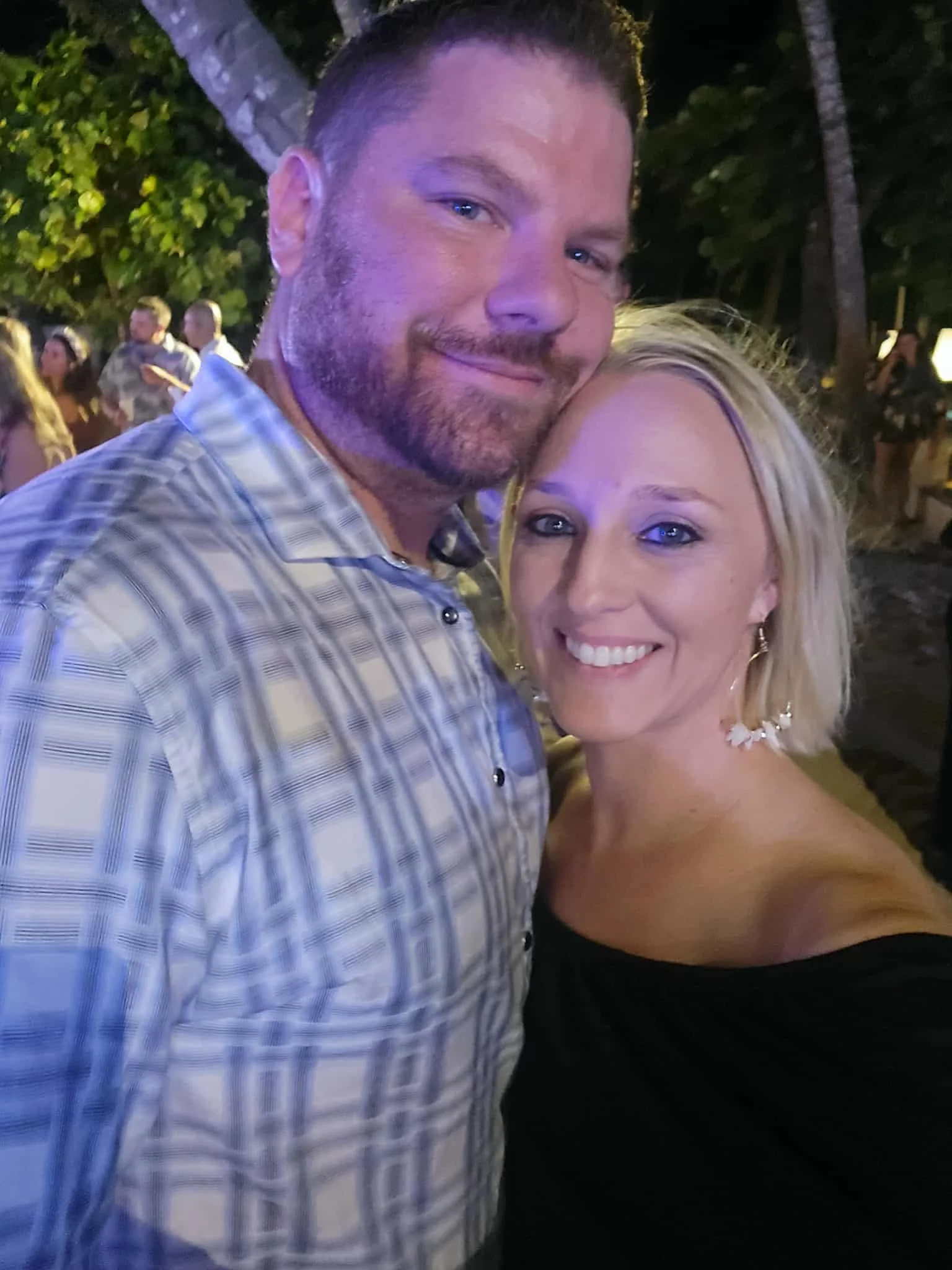 A man and woman smiling for a selfie outdoors at night, with trees and other people in the background.