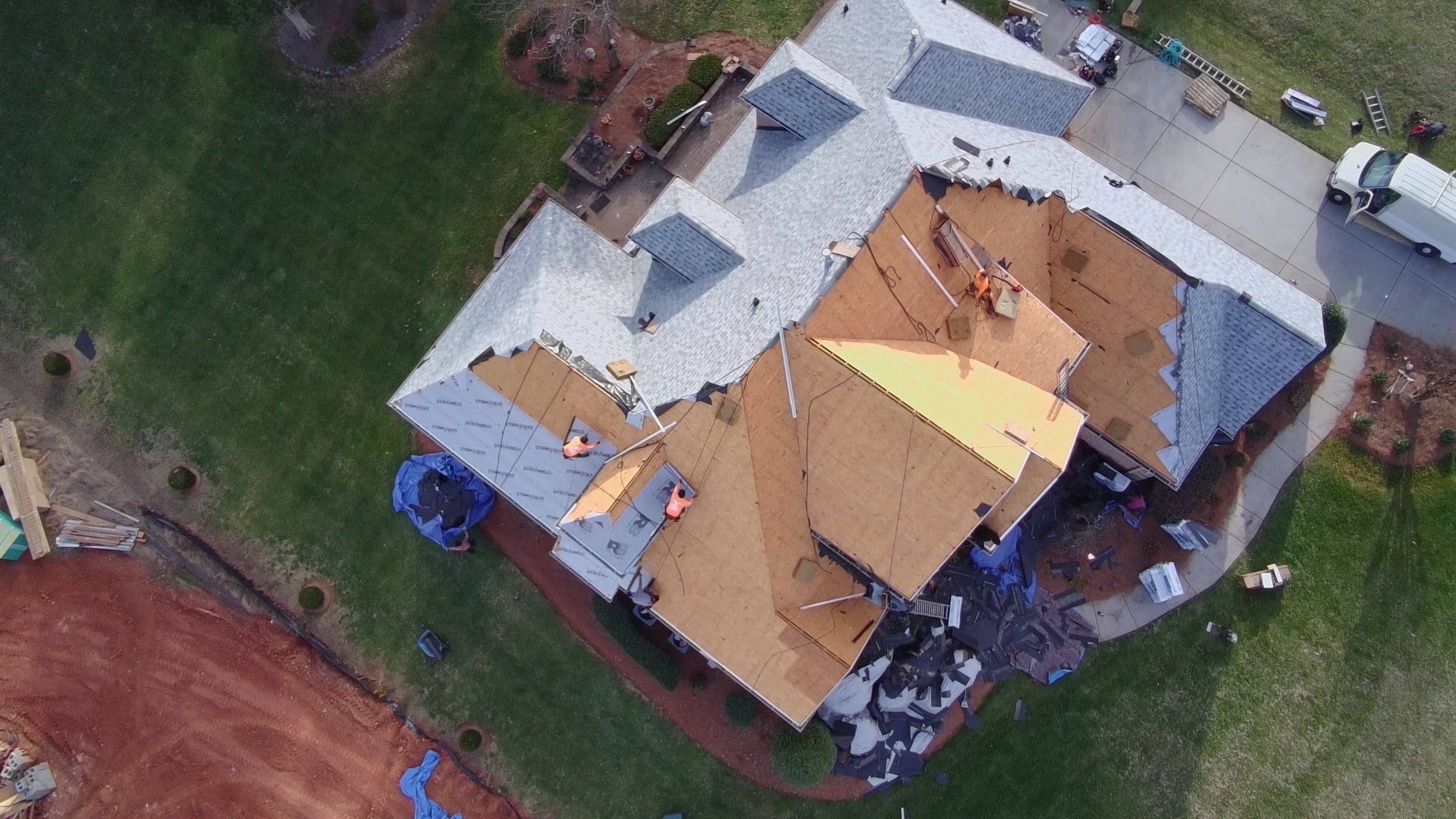 An aerial view of a house under construction with exposed wooden framing, surrounding completed roofing, driveway, and green lawn.