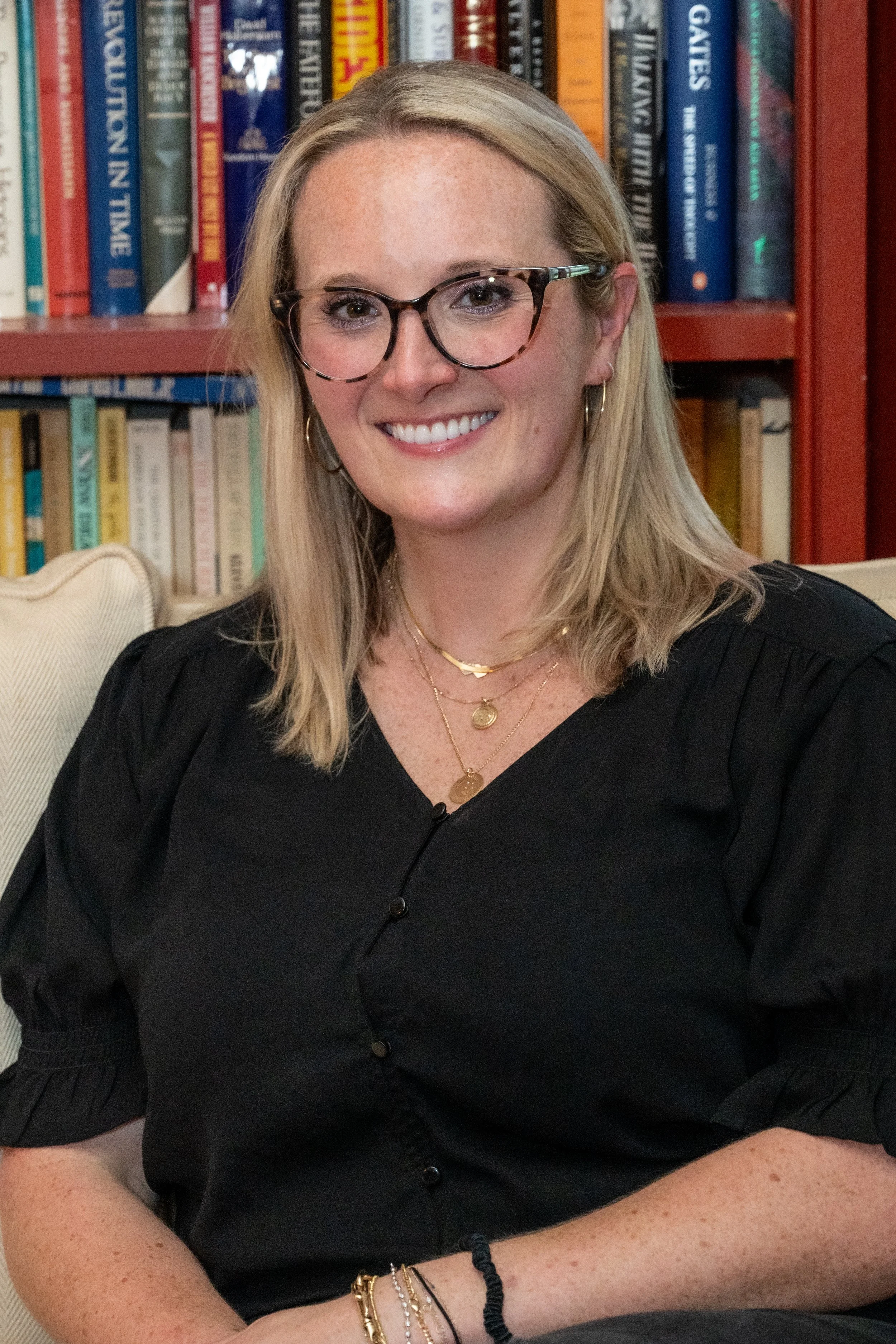 A woman with blonde hair, glasses, and jewelry smiling in front of a bookshelf filled with books.