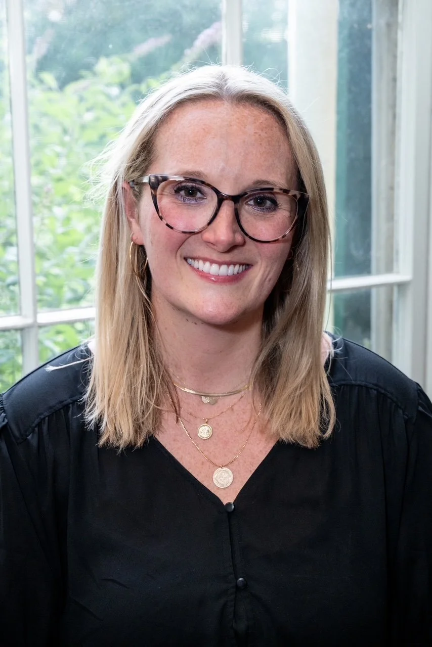 A smiling woman with blonde hair, glasses, and layered gold necklaces, wearing a black top, sitting in front of a window with greenery outside.