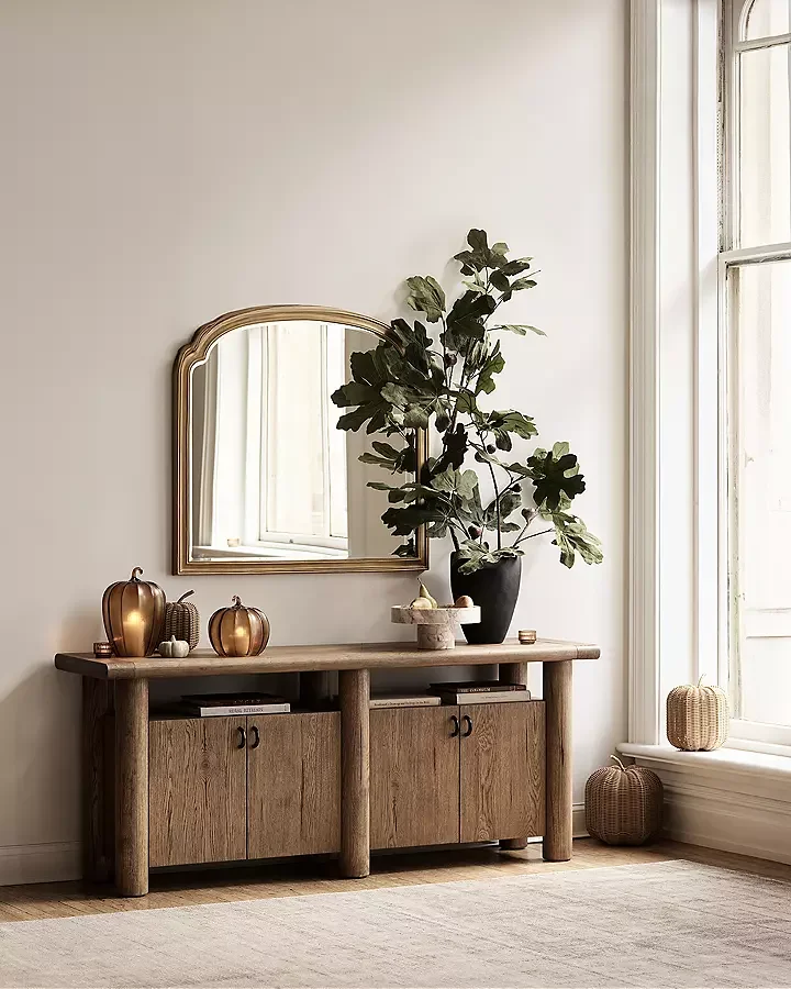 A wooden console table with decor, a mirror, a large potted plant, and wicker baskets near a window in a minimalist living space.