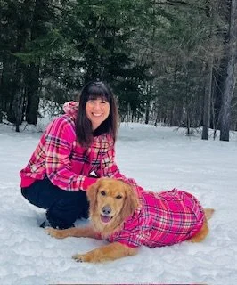 Woman and dog in matching pink plaid outfits sitting on snow in a wooded area