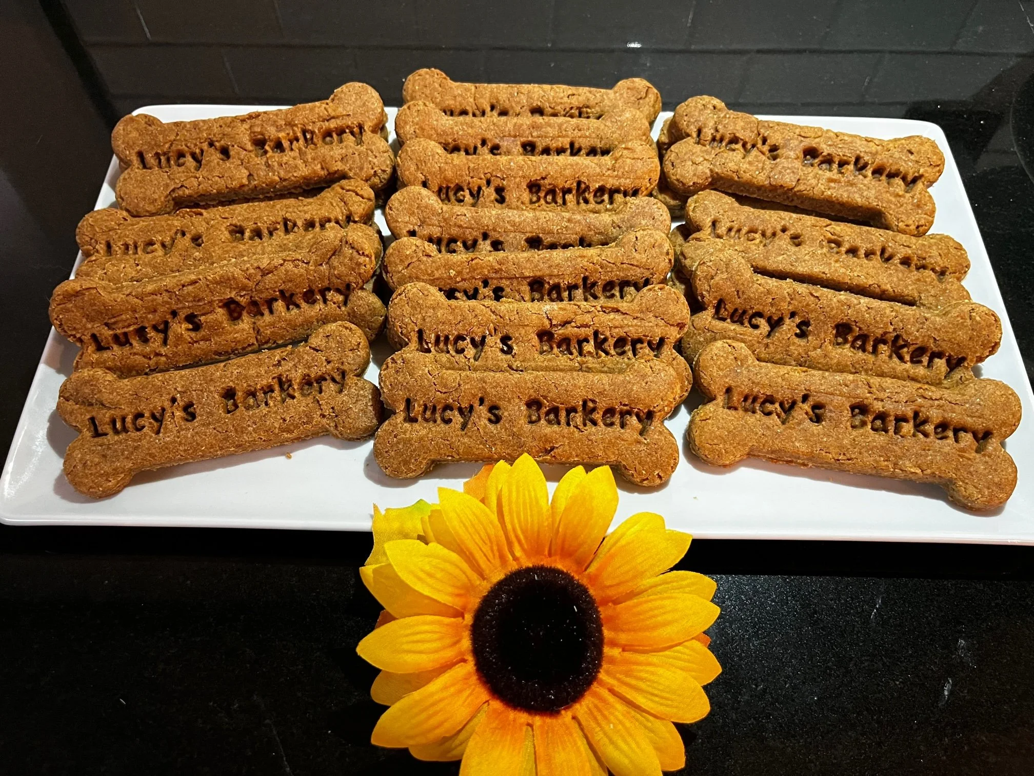 A white rectangular plate displays dog biscuits shaped like bones, each engraved with 'Lucy's Barkery.' A yellow sunflower is placed in front of the plate on a black surface.