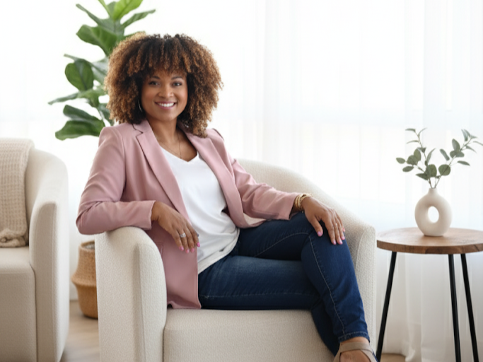 Keisha Gaddis sitting on a cream-colored armchair in a bright room with plants and a side table.