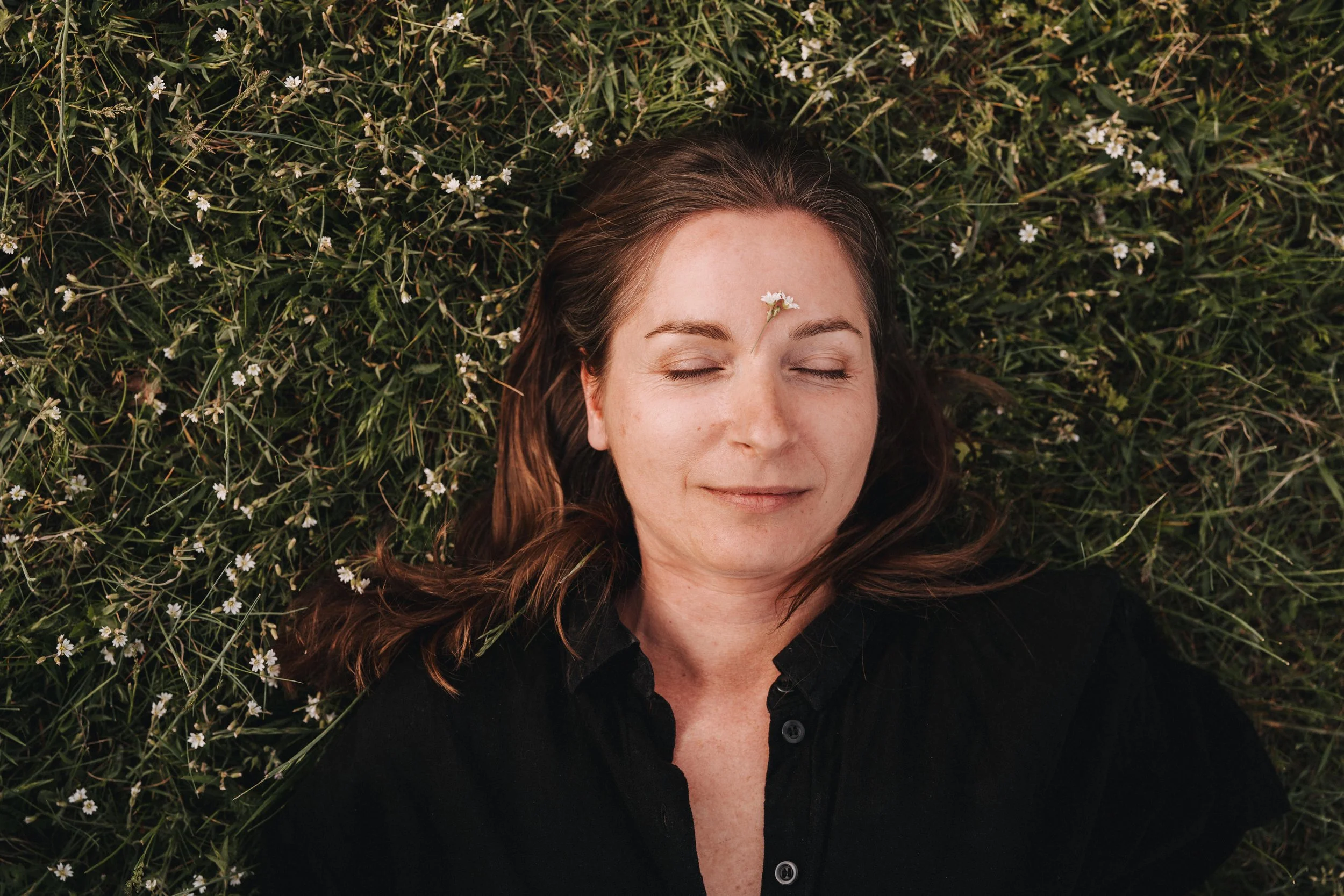 Frau mit geschlossen Augen liegt auf einer Wiese mit kleinen weißen Blumen, ein kleines Blumenstück liegt auf ihrer Stirn.