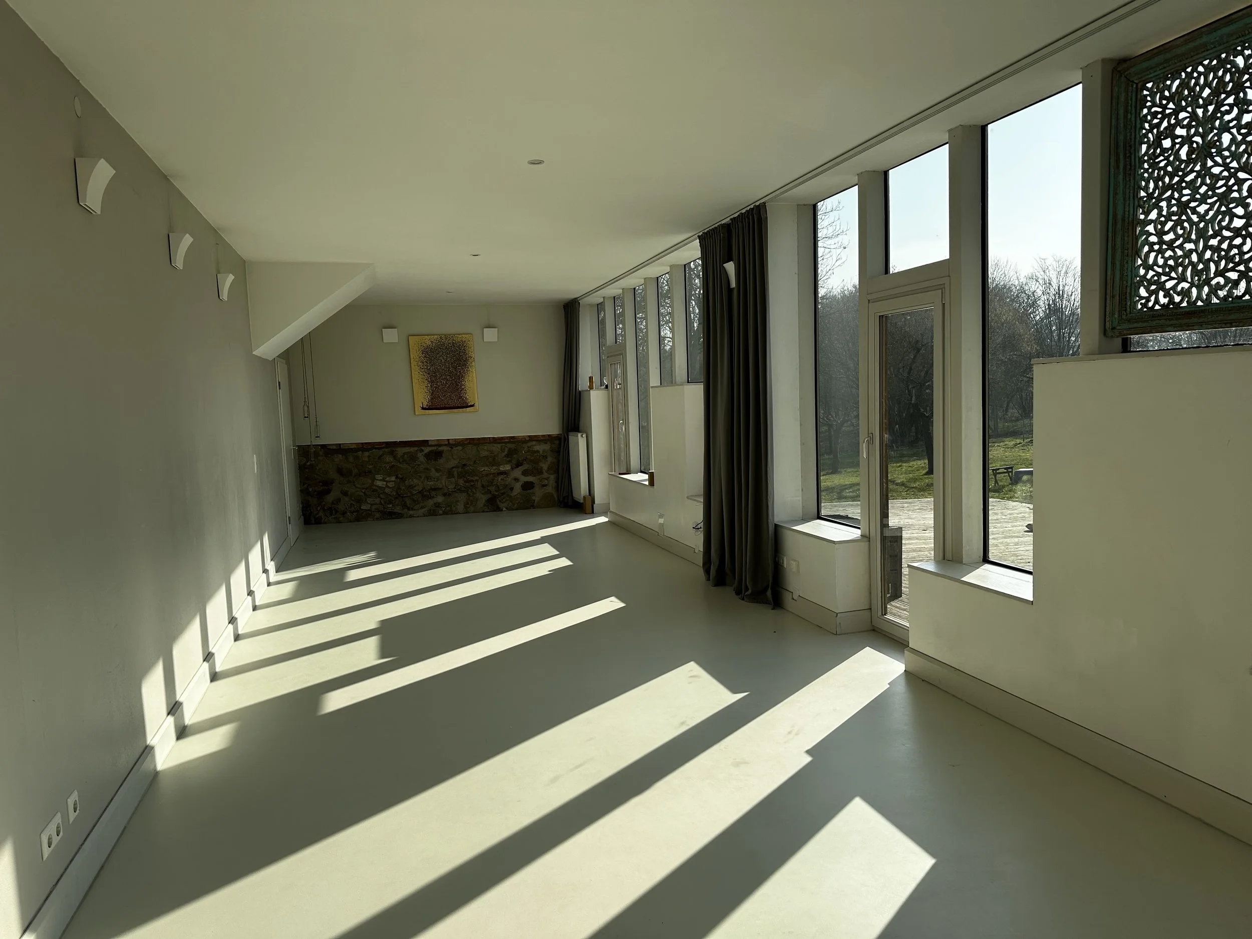 Leeres helles Zimmer mit großen Fenstern, durch die Sonnenlicht und Schatten auf den Boden fallen.