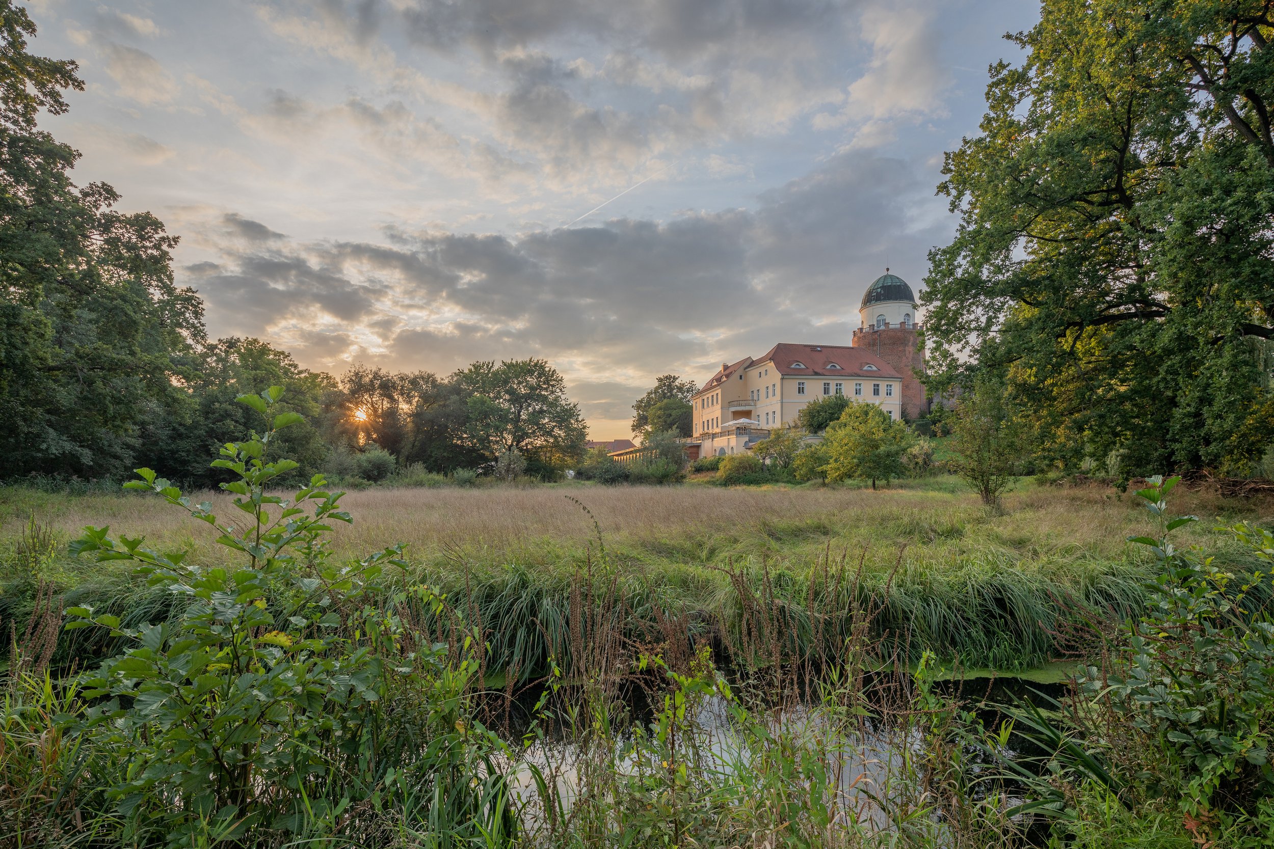 Ein idyllischer Blick auf eine grüne Wiese mit einem kleinen Wasserlauf im Vordergrund. Im Hintergrund sieht man ein Gebäude mit roten Dächern und einer grünen Kuppel, umgeben von Bäumen bei Sonnenuntergang, mit einem bewölkten Himmel.