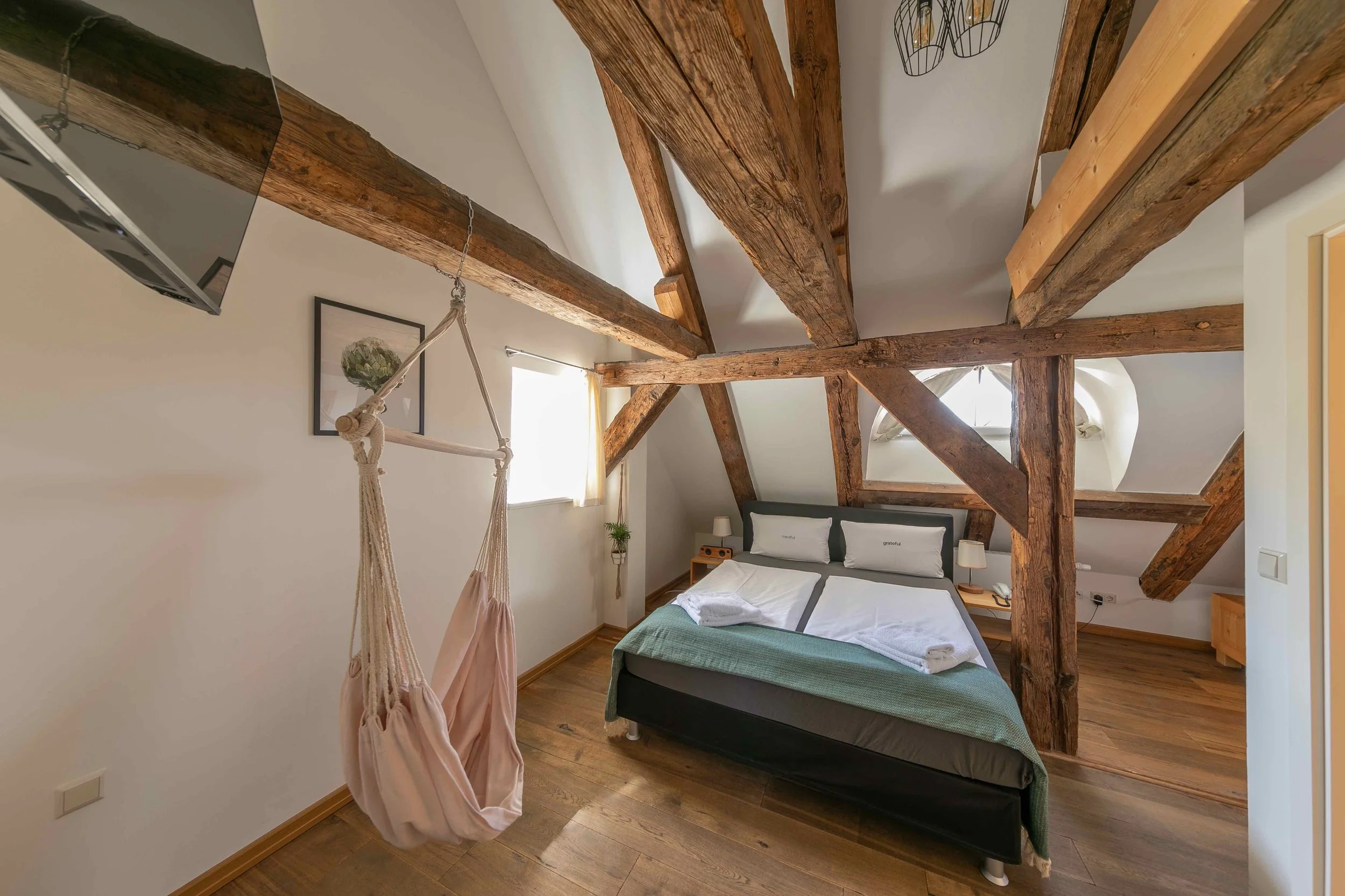 Ein Schlafzimmer mit einem Doppelbett, Holzdeckenbalken und einem kleinen Fenster an der Decke. Daneben hängt eine Hängehöhle an der Decke, und auf den Nachttischen stehen Lampen. Die Wände sind weiß, und der Boden ist holzfarben.
