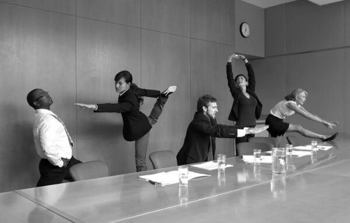 Fünf Menschen im Anzug zeigen verschiedene akrobatische Posen in einem Konferenzraum, während sie um den Tisch sitzen.